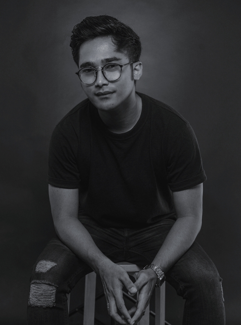 Black and white half-body shot of Raimi Safari seating on a stool, wearing glasses, fingers lightly pressed together.