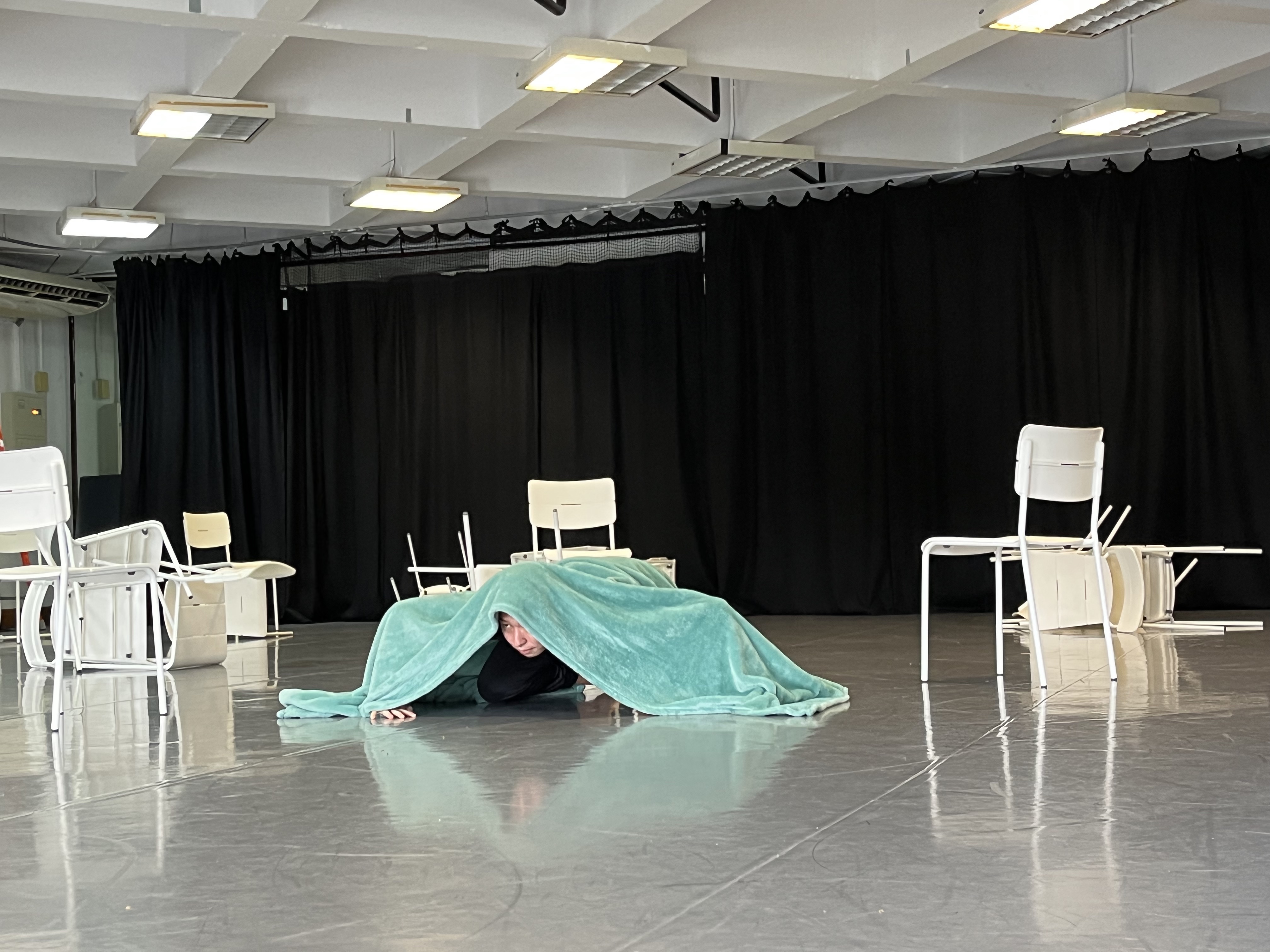 An image of Chng Yi Kai crouched under a large green cloth surrounded by white chairs in disarray.