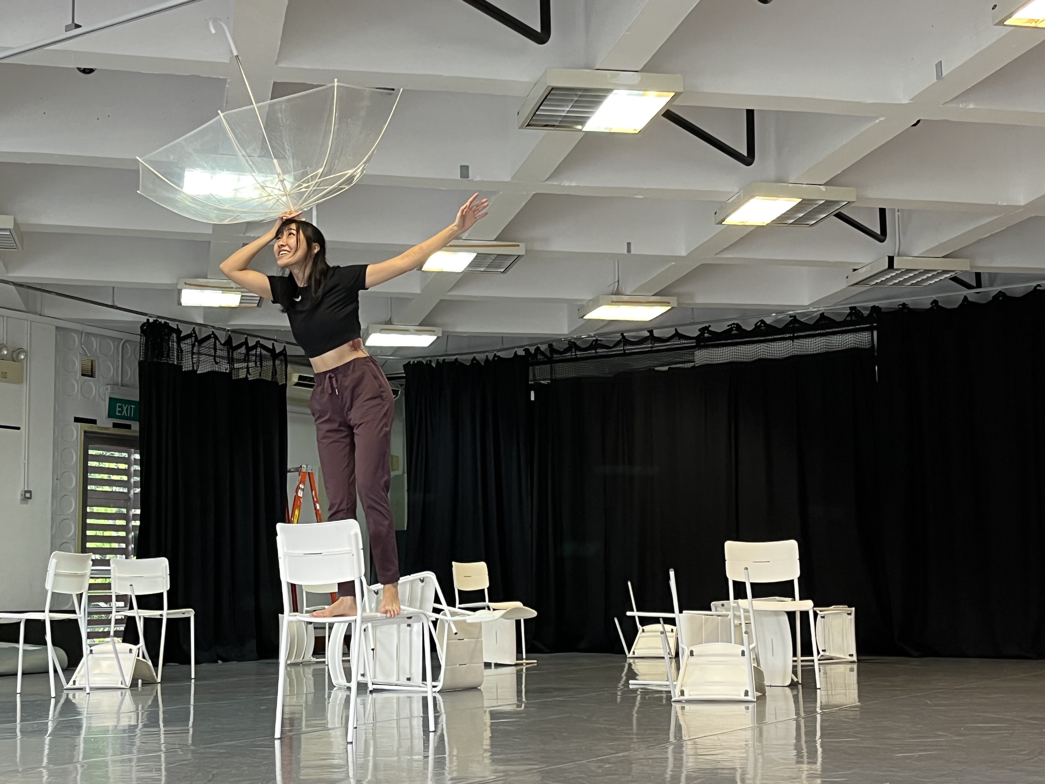 An image of Ella Wee standing atop a white chair, arms raised and holding an upturned transparent umbrella on her head.