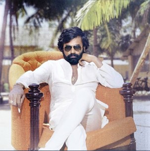 Bada Jabari sporting sunglasses and wearing a white jumpsuit while seated on a chair cross-legged, staring directly at the camera.