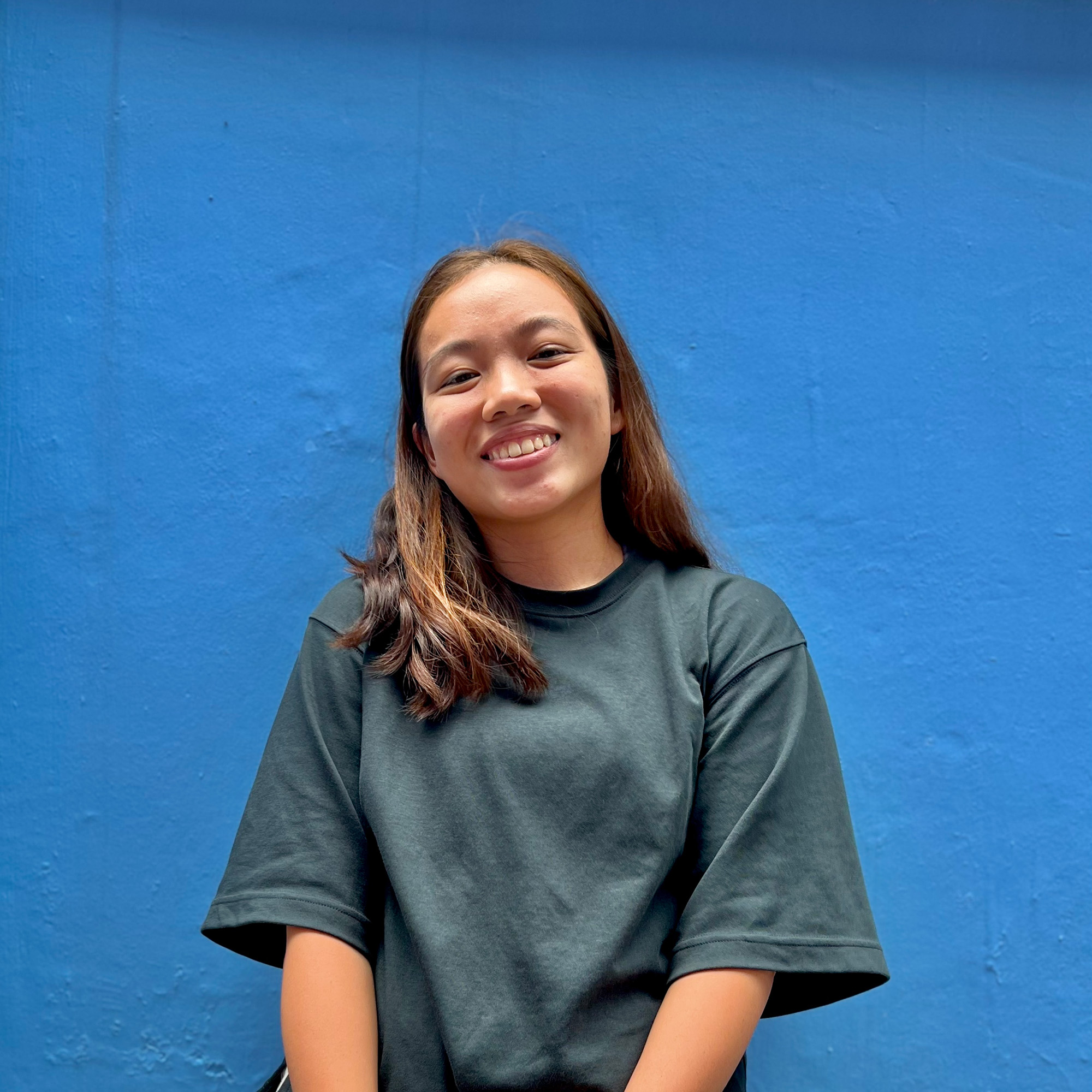 Half-body shot of Dylia Ang in a dark grey shirt, against a blue wall, smiling down at the camera.