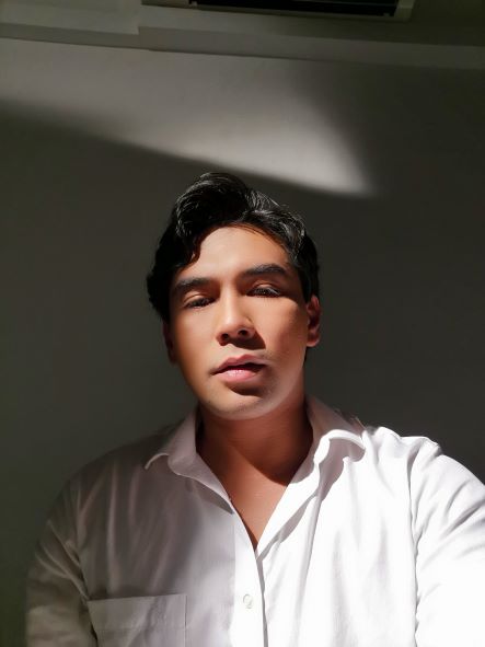 Irfan Kasban wearing a white shirt looking down at the camera in room with strong shadows behind him.