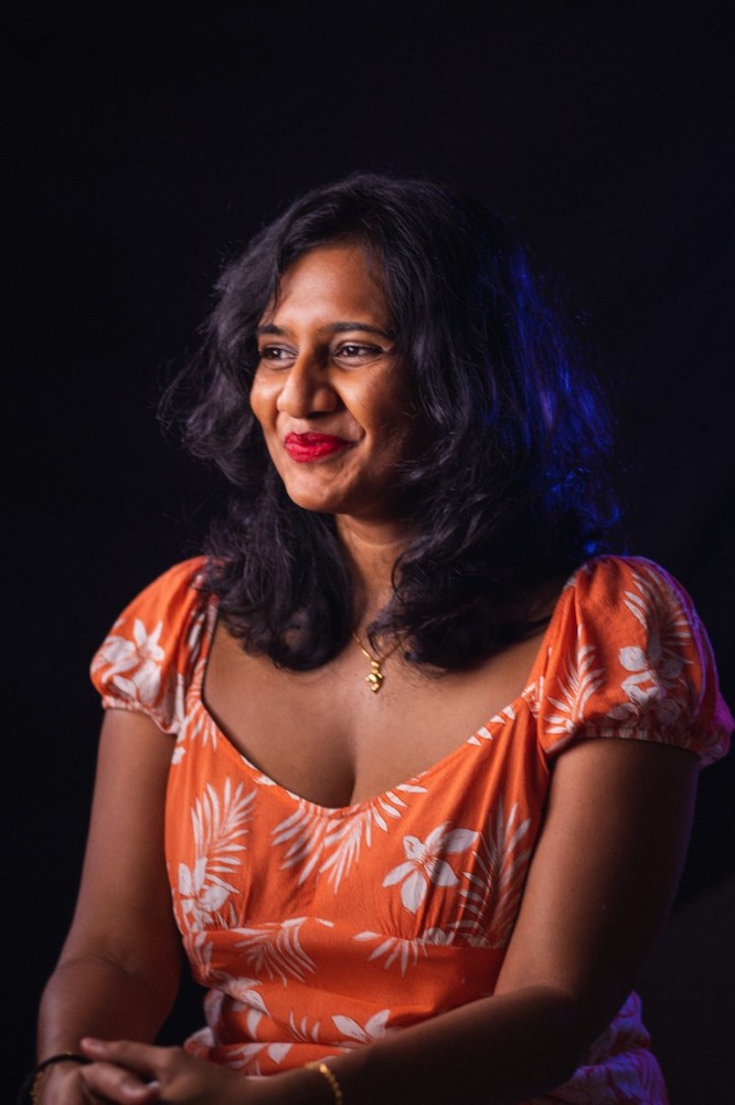 Half-body shot of Melizarani T Selva in an orange dress, seated with hands clasped, smiling and glancing away from the camera.