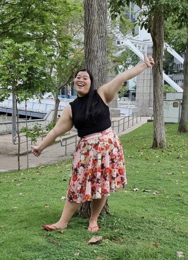 Full-body shot of Miriam Cheong posing and smiling in front of a tree, wearing a sleeveless black top and colourful skirt.