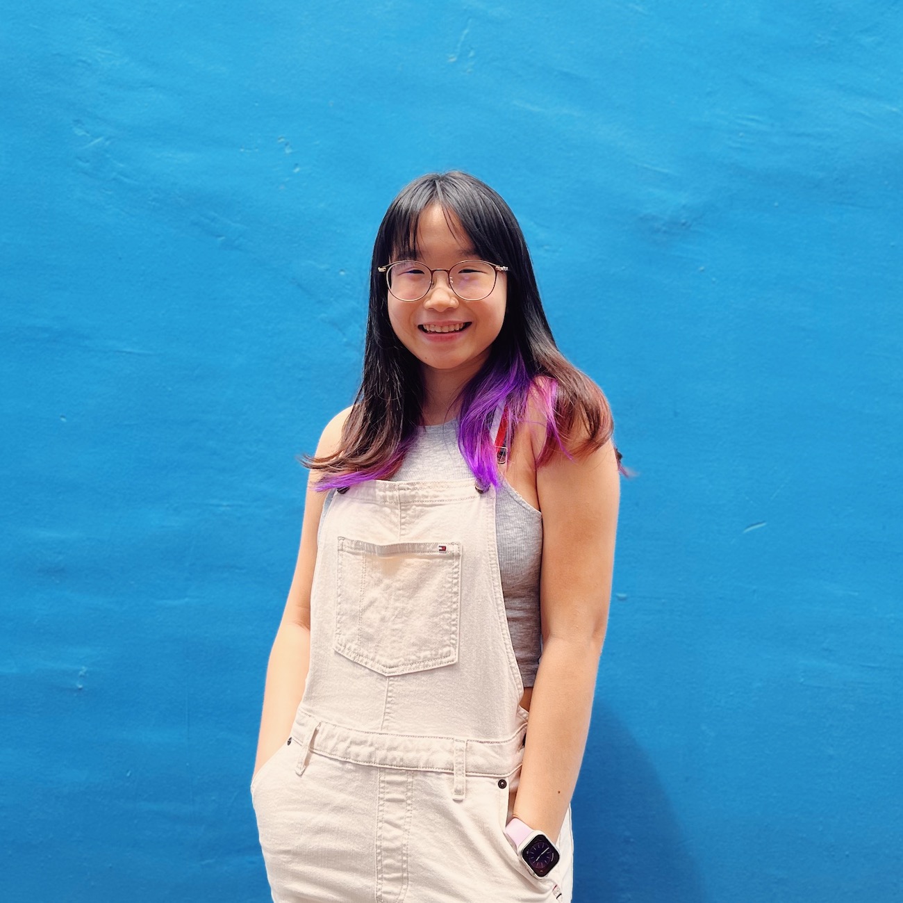 Half-body shot of Voon Yue Qi in white overalls against a blue wall, smiling with her hands in her pockets.