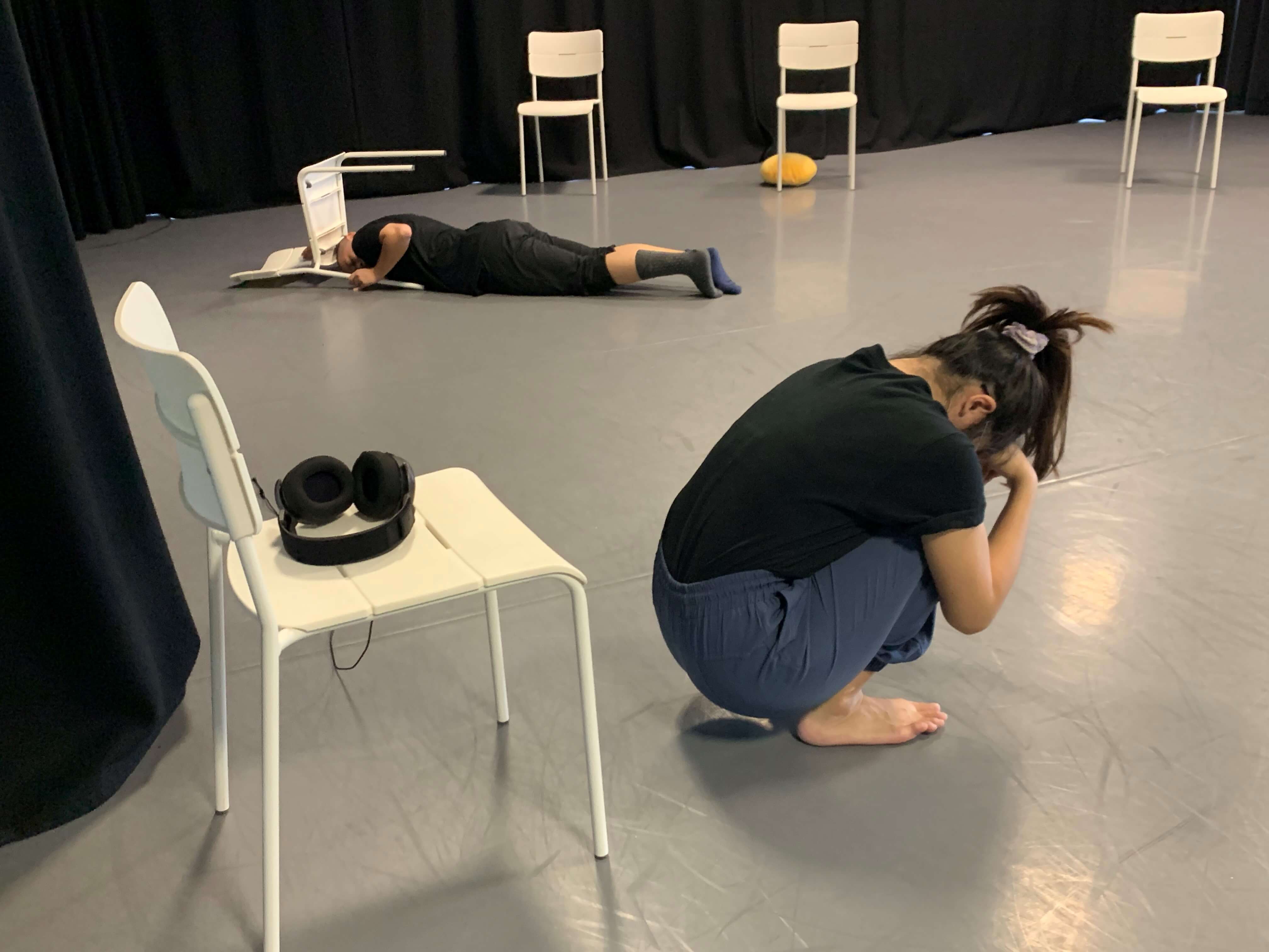 A photograph of a rehearsal where a female presenting person is squatting in front of a chair with her head in her hands and another female presenting person in the background lying on the floor next to a chair.