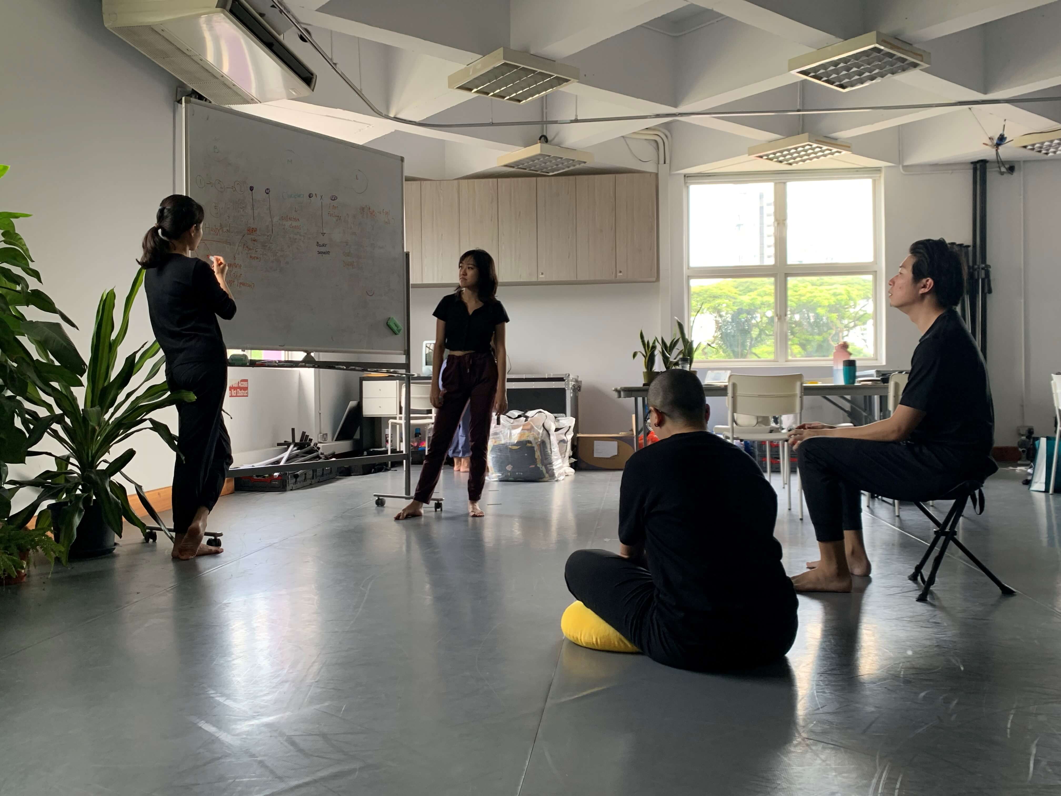 Photograph of a discussion where two female presenting people dressed in black shirt and pants stand next to a whiteboard with notes, while one person sits on the floor and another sitting on a chair listening in to the discussion.