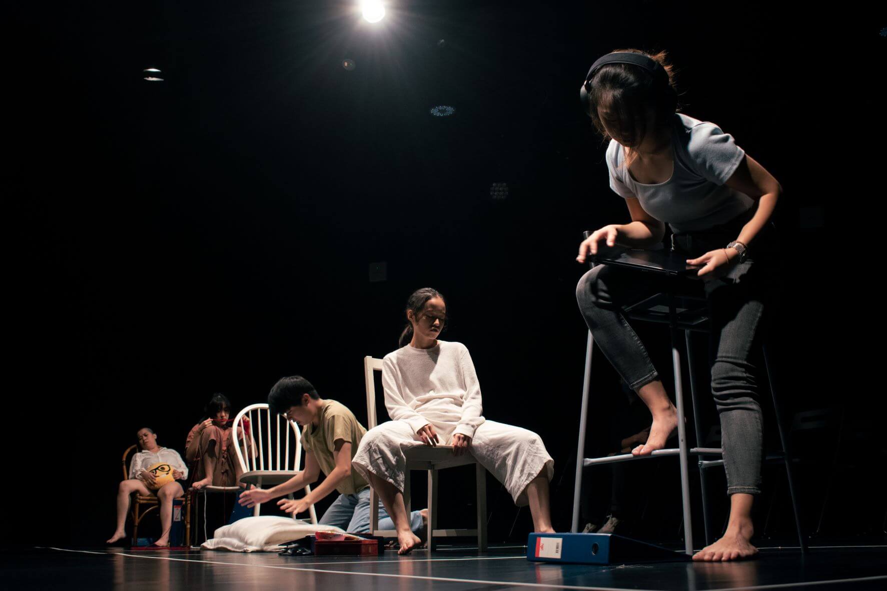 The photograph features a still from a performance in a black box where five people are each seated on or near their chair in various poses expressing ennui. 