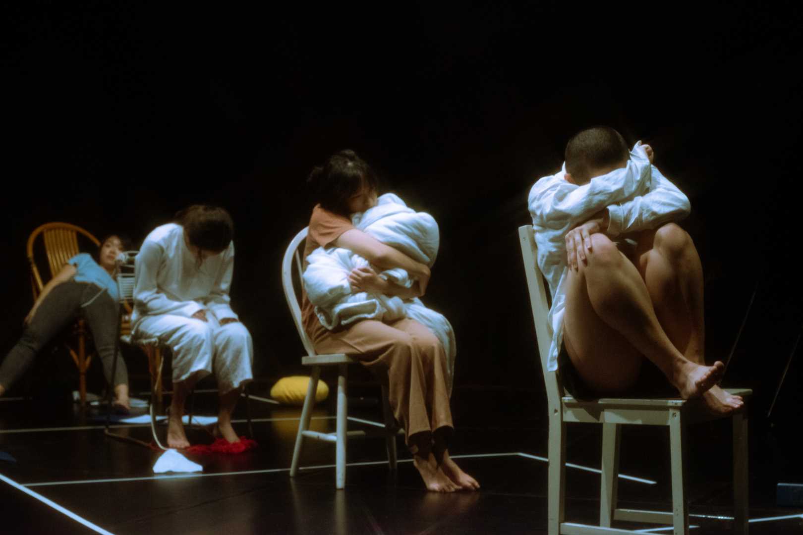 The photograph features a still from a performance in a black box where four performers are seated on chairs, the first performer closest to the camera is hugging her knees on the chair, the second is hugging a blanket, the third is bowed over, and the last performer is slumped over her chair.