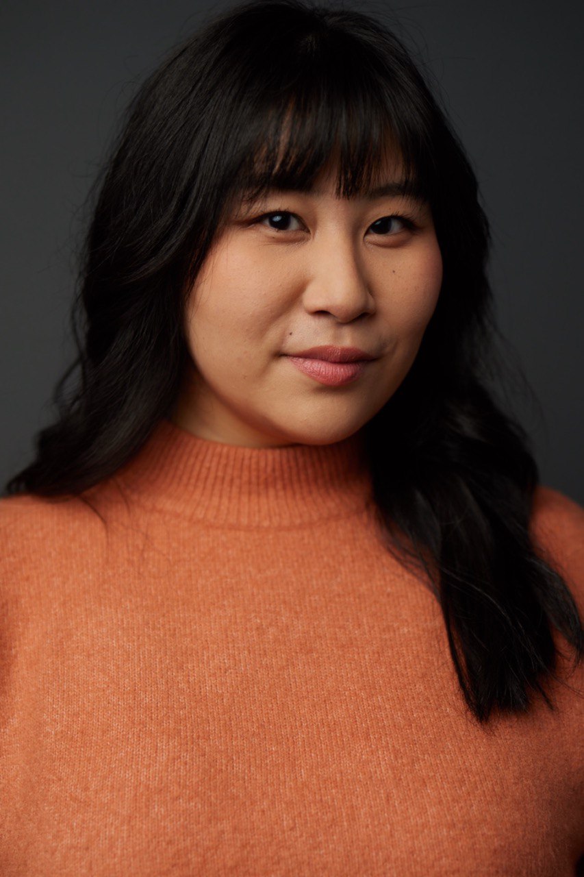 Headshot of Ashley Ru in an orange turtleneck, with long hair.