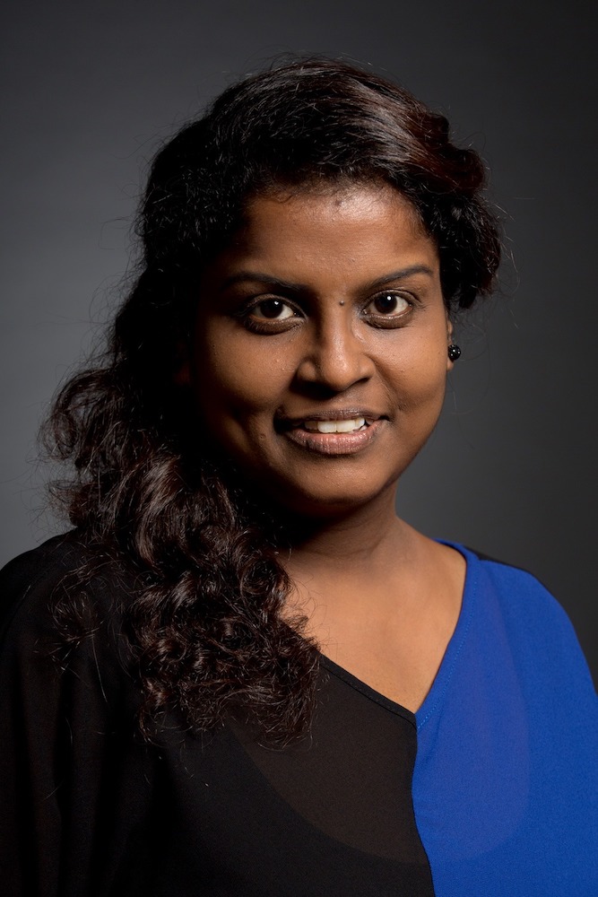 Half-body shot of Grace Kalaiselvi in a v-neck shirt and long curly hair, smiling.