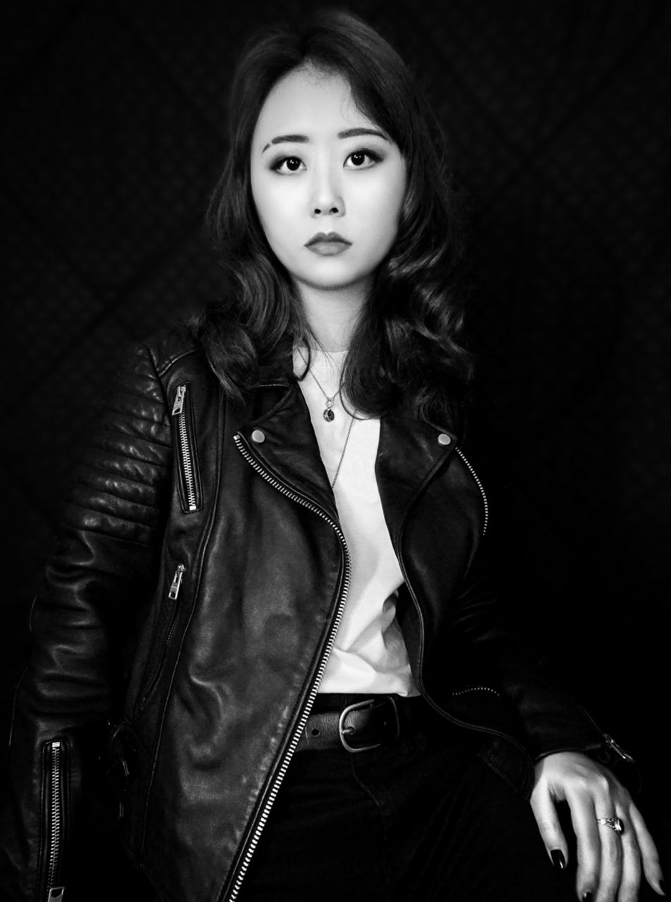 Half-body black and white shot of Juniper Lee in a leather jacket and white shirt, staring into the camera.