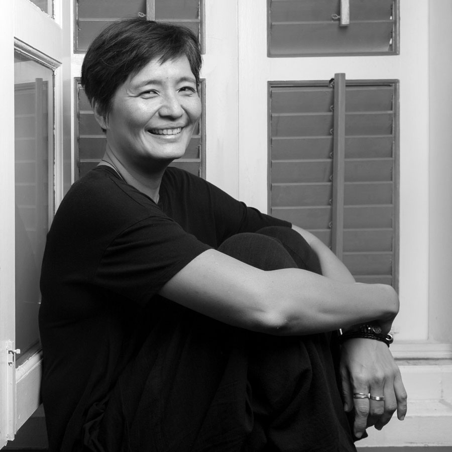 Black and white shot of Kuo Jian Hong seated on the floor in front of window shutters, smiling with her arms around her knees.