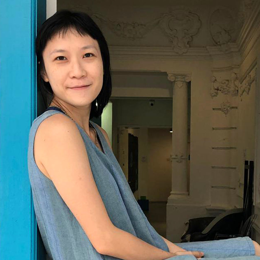 Half-body shot of Ng Mun Poh seated in a window frame in front of a building, hands in her lap, smiling.