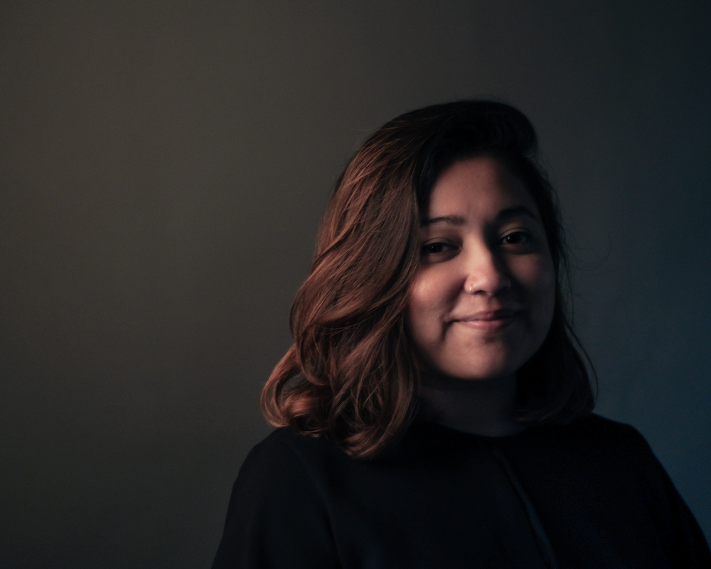 Coloured headshot of Shaza Ishak, in a dark coloured shirt against a dark grey background.