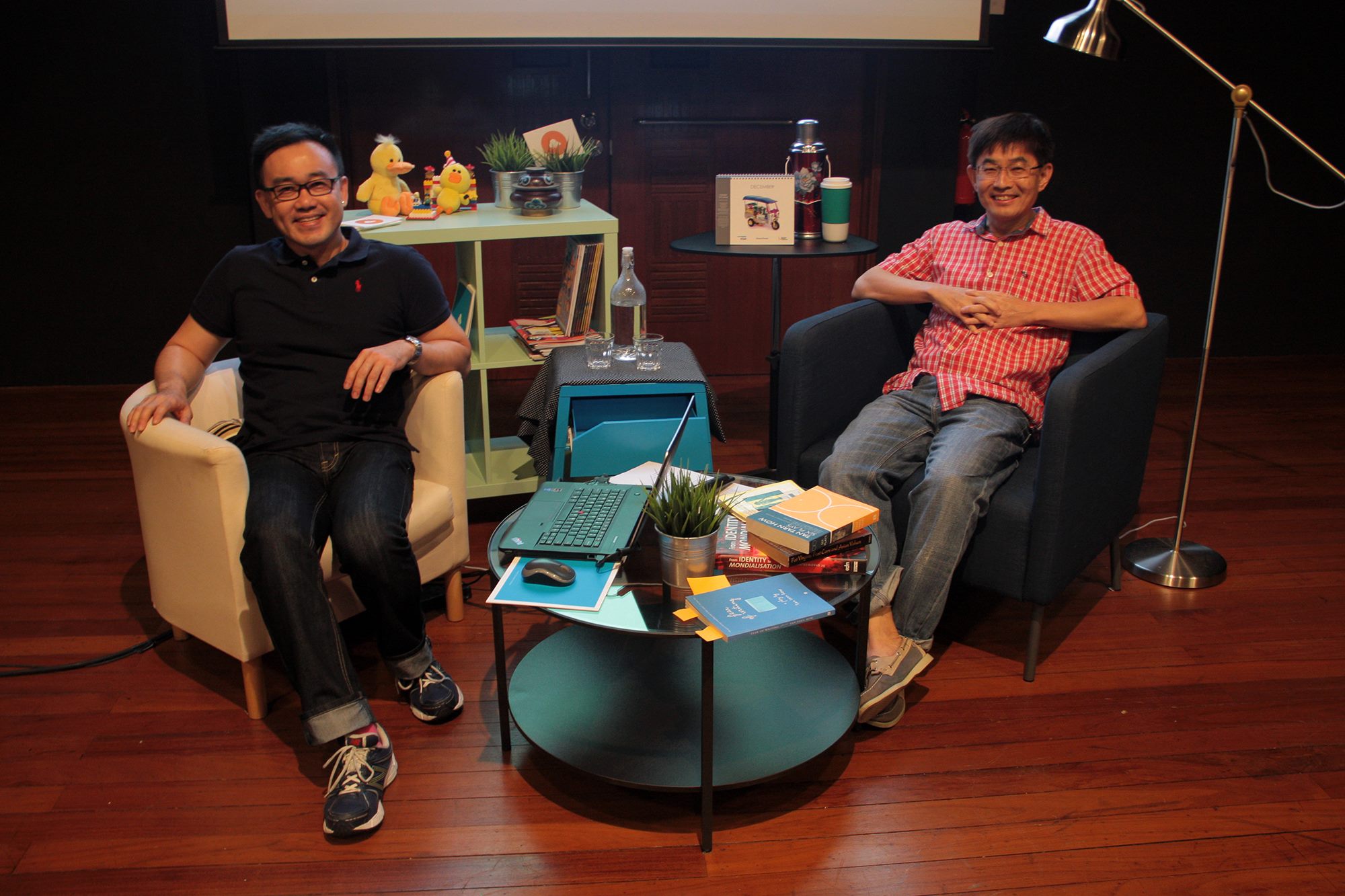 The photograph features Robin Loon on the left and Tan Tarn How on the right. Both are seated in chairs, with multiple objects including books, a laptop and decorations giving the sense of a living room.