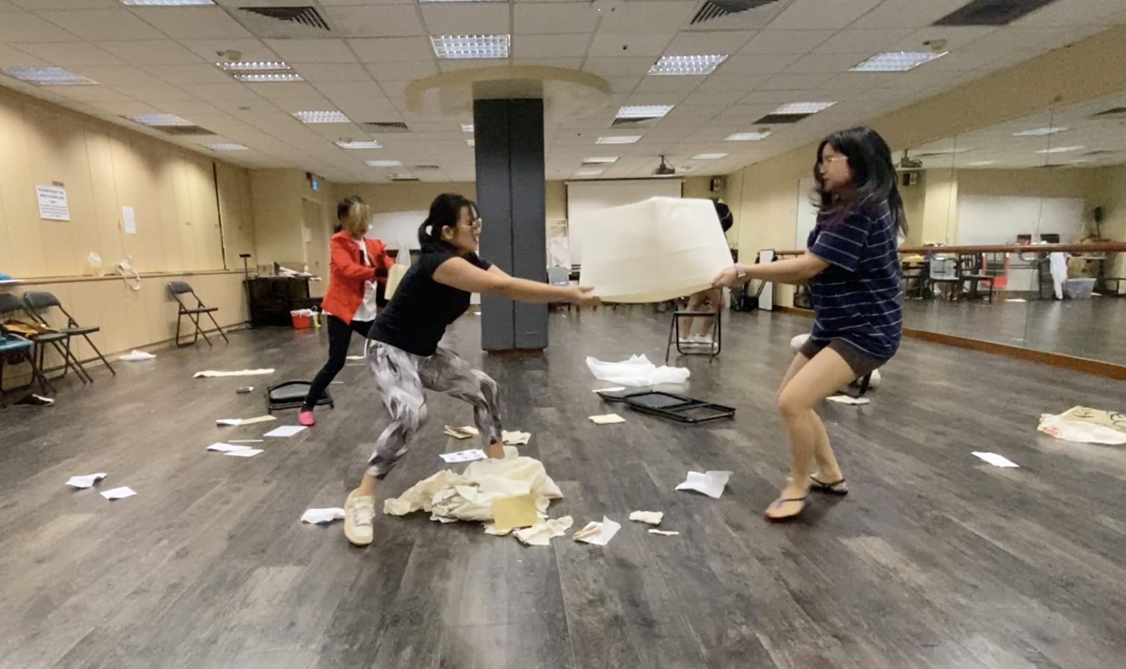 The photograph of a rehearsal featuring two female presenting people tugging a white box between themselves. A person wearing a red sweater is behind them. Paper is littered over the floor.