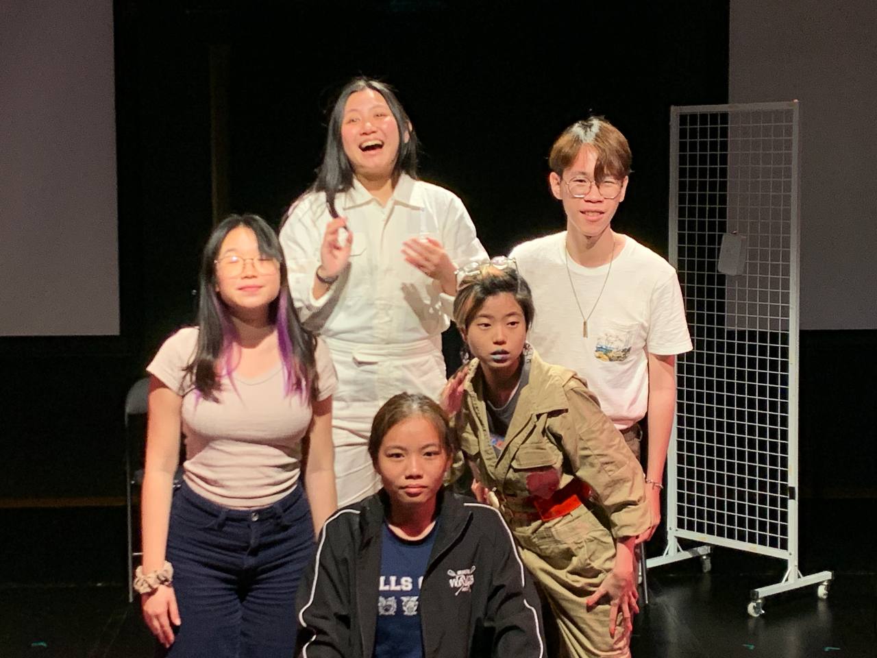 The photograph features a still from a rehearsal in a black box where Ariel Lim, Voon Yueqi, Noah Lee, Michael Neo, and Dylia Ang are standing together, displaying various expression from laughter to stoic gazes.