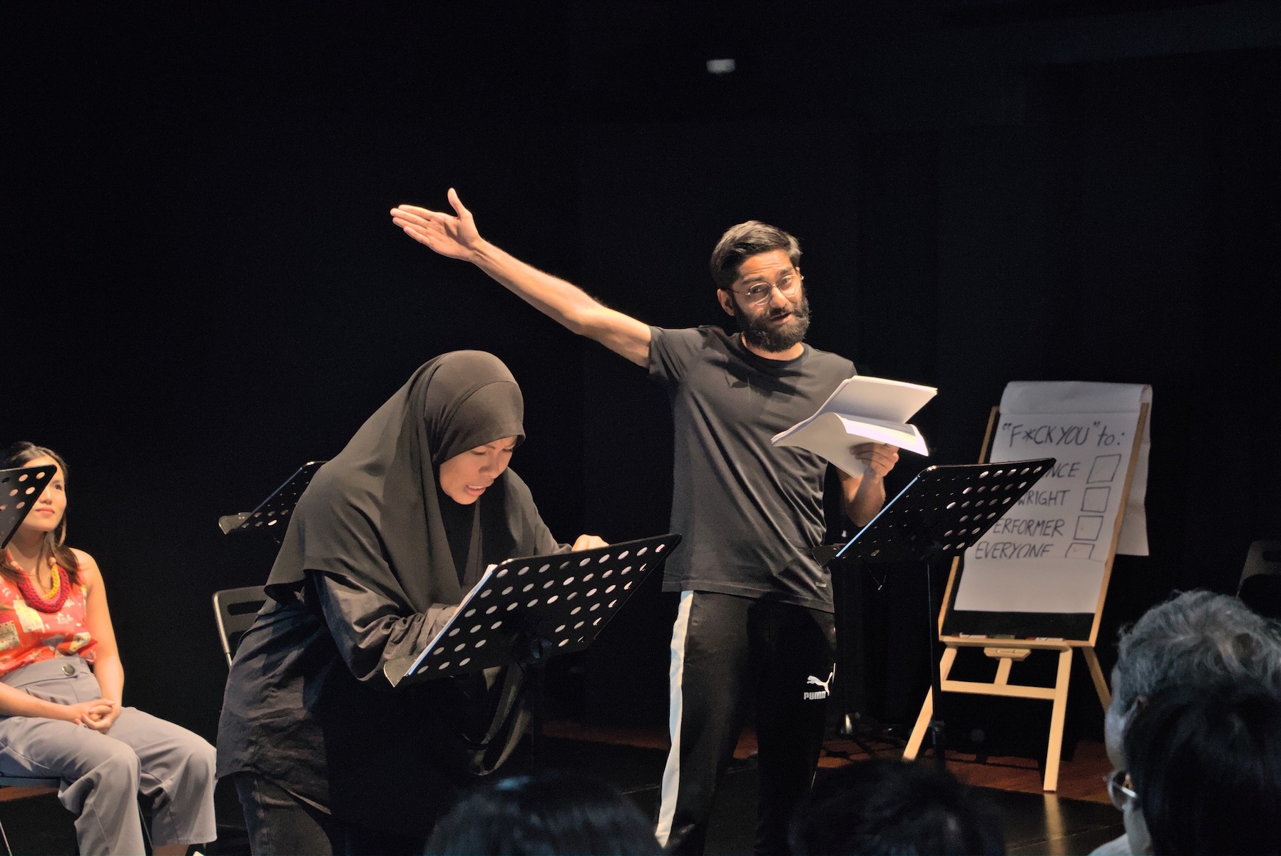 A male-presenting person gesturing widely with his right hand. Beside him, a distressed-looking female-presenting person in a hijab bends over a script.