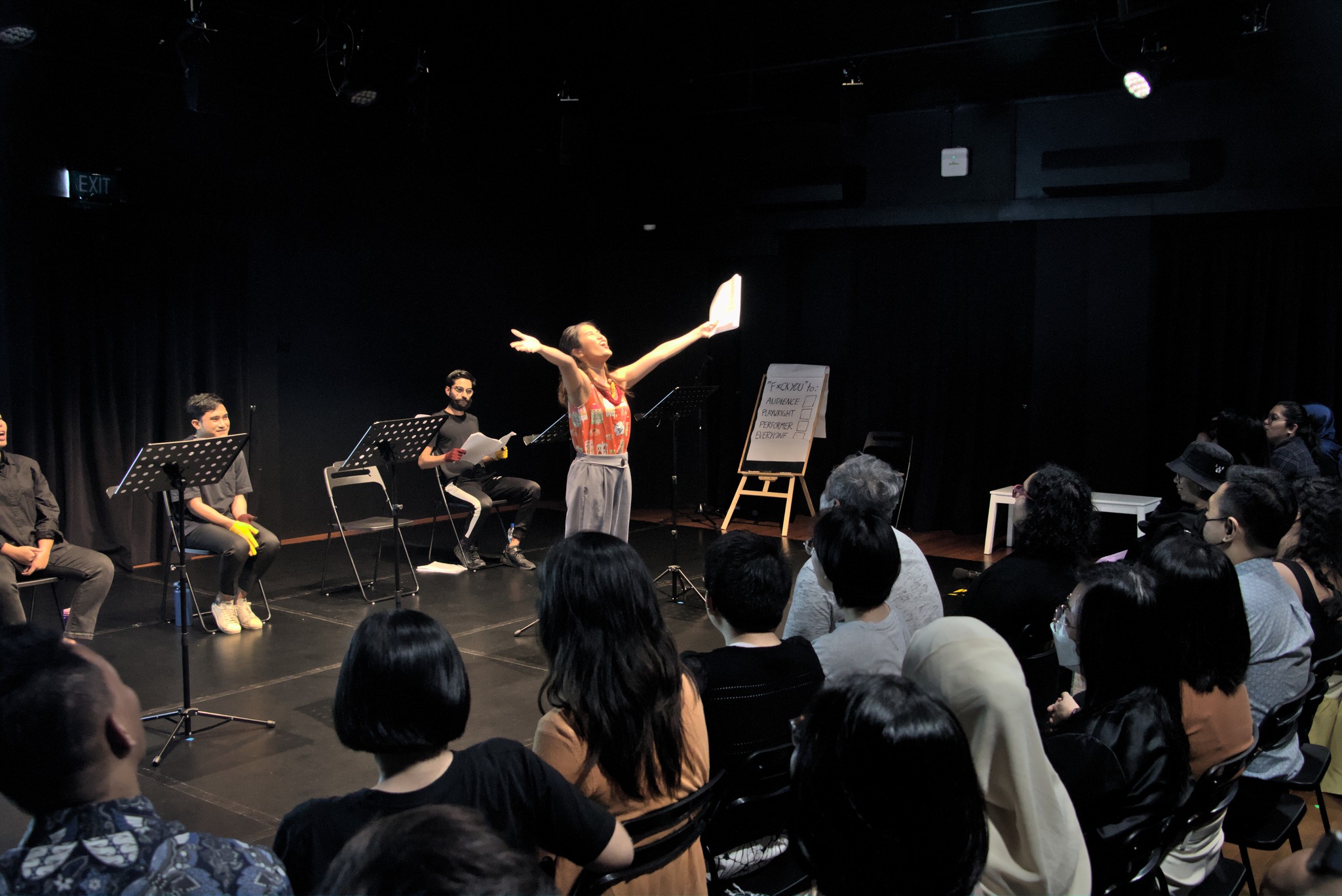 A female-presenting person standing in front of a large audience with her arms outstretched, face up, shouting. She is holding a piece of paper in her left hand.