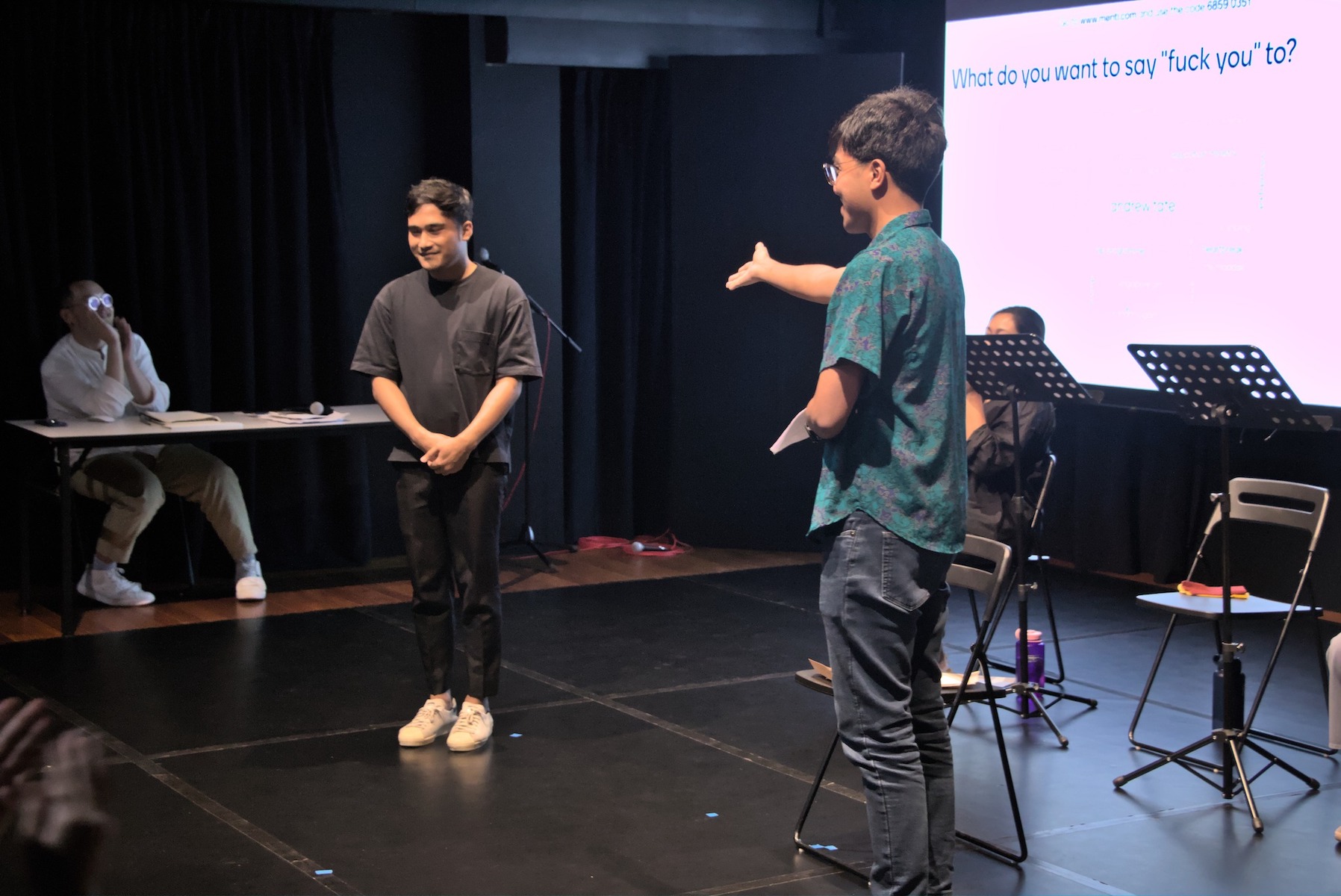 A male-presenting person in a green shirt, right hand outstretched towards another male-presenting person a short distance away, hands clasped in front of him and smiling.