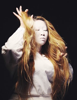 Half-body shot of Gloria Tan with long blonde hair and white face makeup, eyes closed, fingers running through her hair.