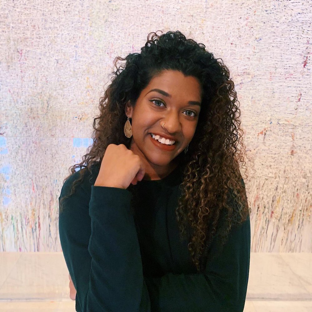 Half-body shot of Indumathi Tamilselvan in front of a white wall, wearing a long-sleeved black shirt. She is smiling, with her fist raised near her chin.