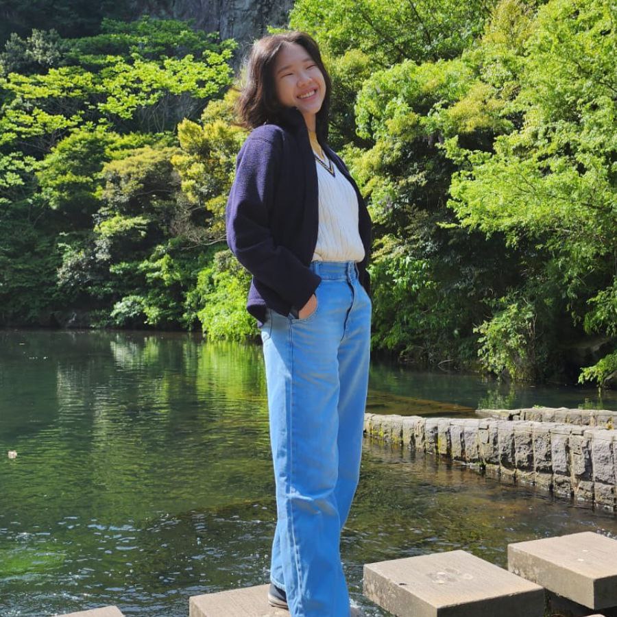 Full-body shot of Letitia Chen by a river lined with lush greenery, standing on stone steps with her hands in her pockets, smiling.