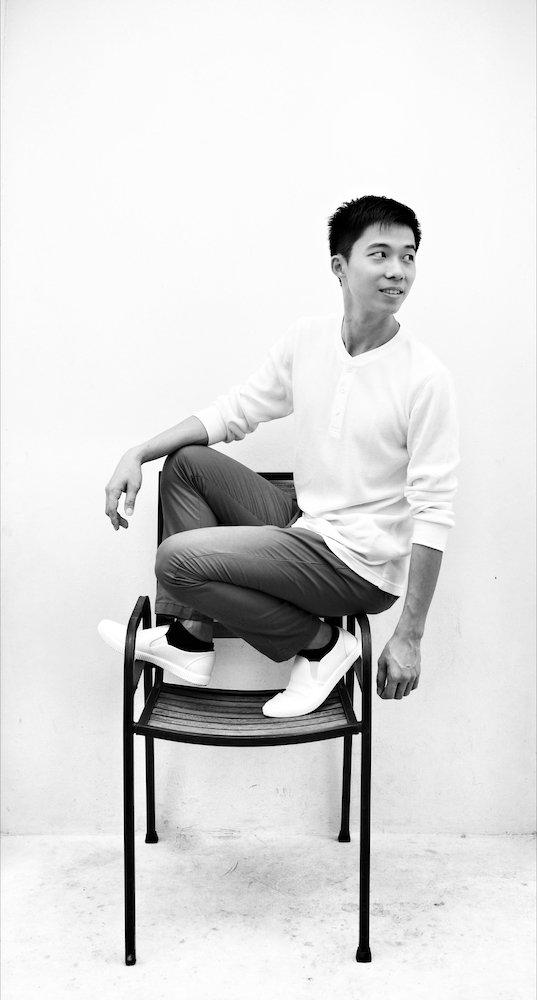 Black and white full-body posed candid shot of Neo Hai Bin, elegantly posed in a chair and glancing to the left, smiling.