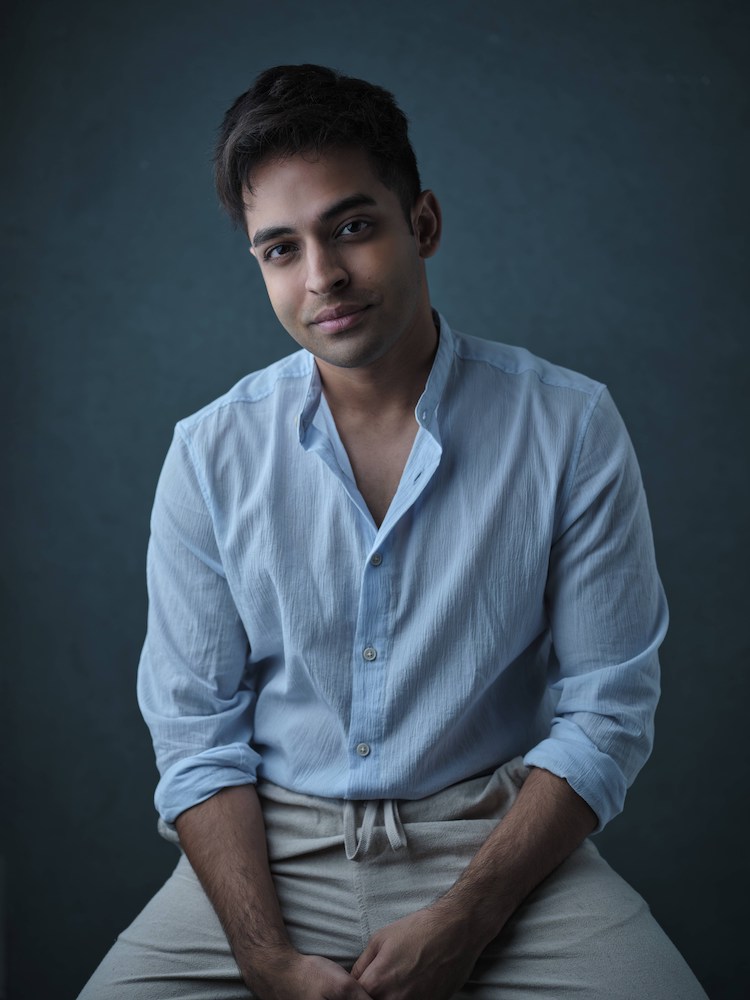 Half-body shot of Shrey Bhargava in a blue shirt and beige pants, seated, hands clapsed, looking into the camera.