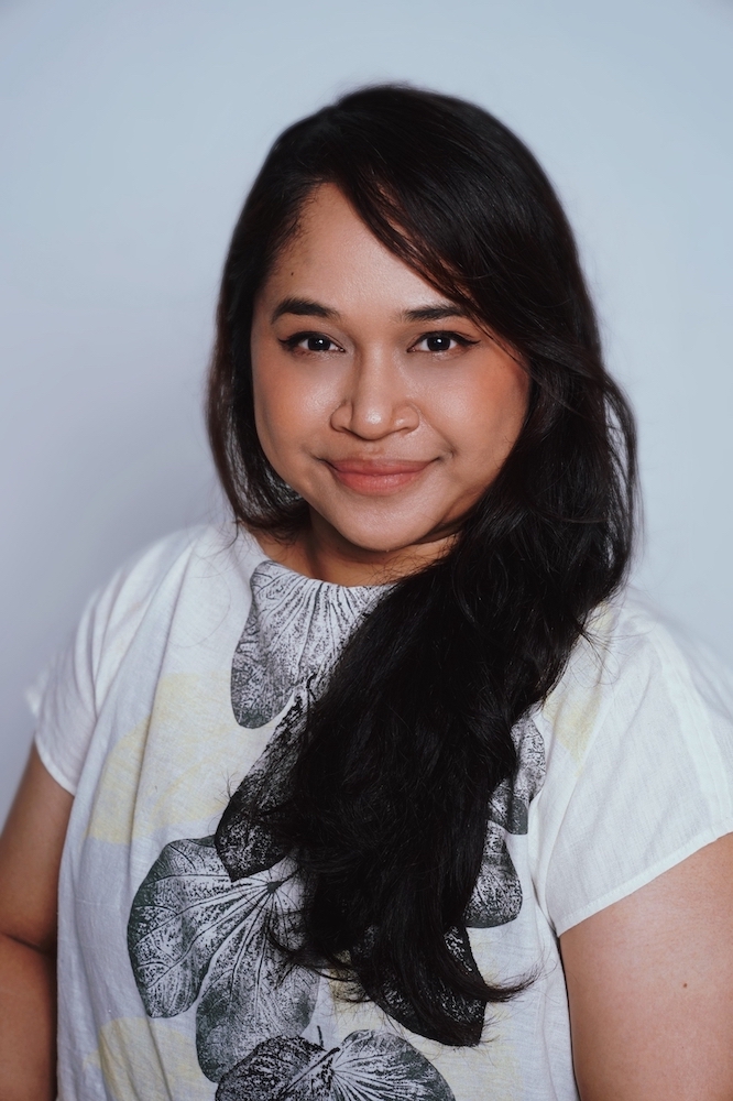 Half-body shot of Suhaili Safari, with long hair, wearing a white top, smiling at the camera.