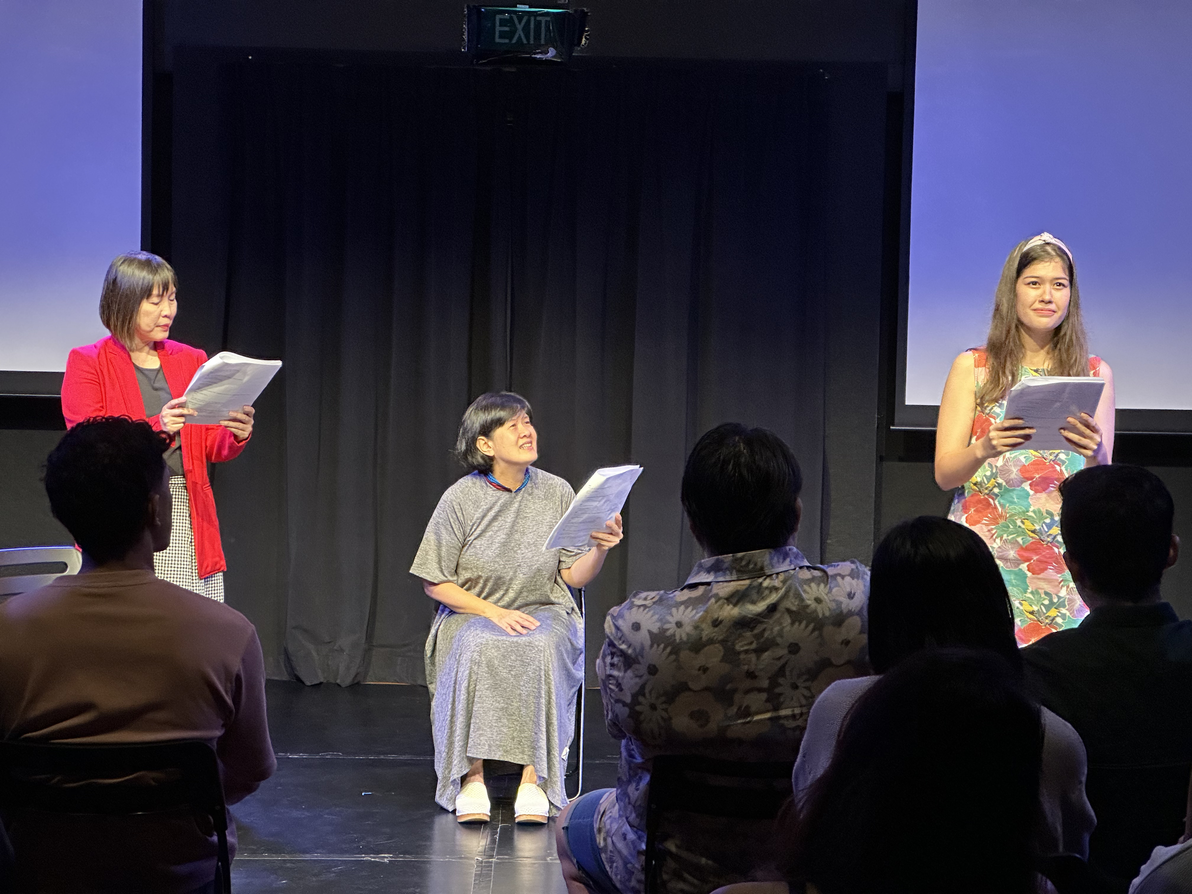 A photograph featuring three actresses, two standing and one seated, each holding a script in hand.