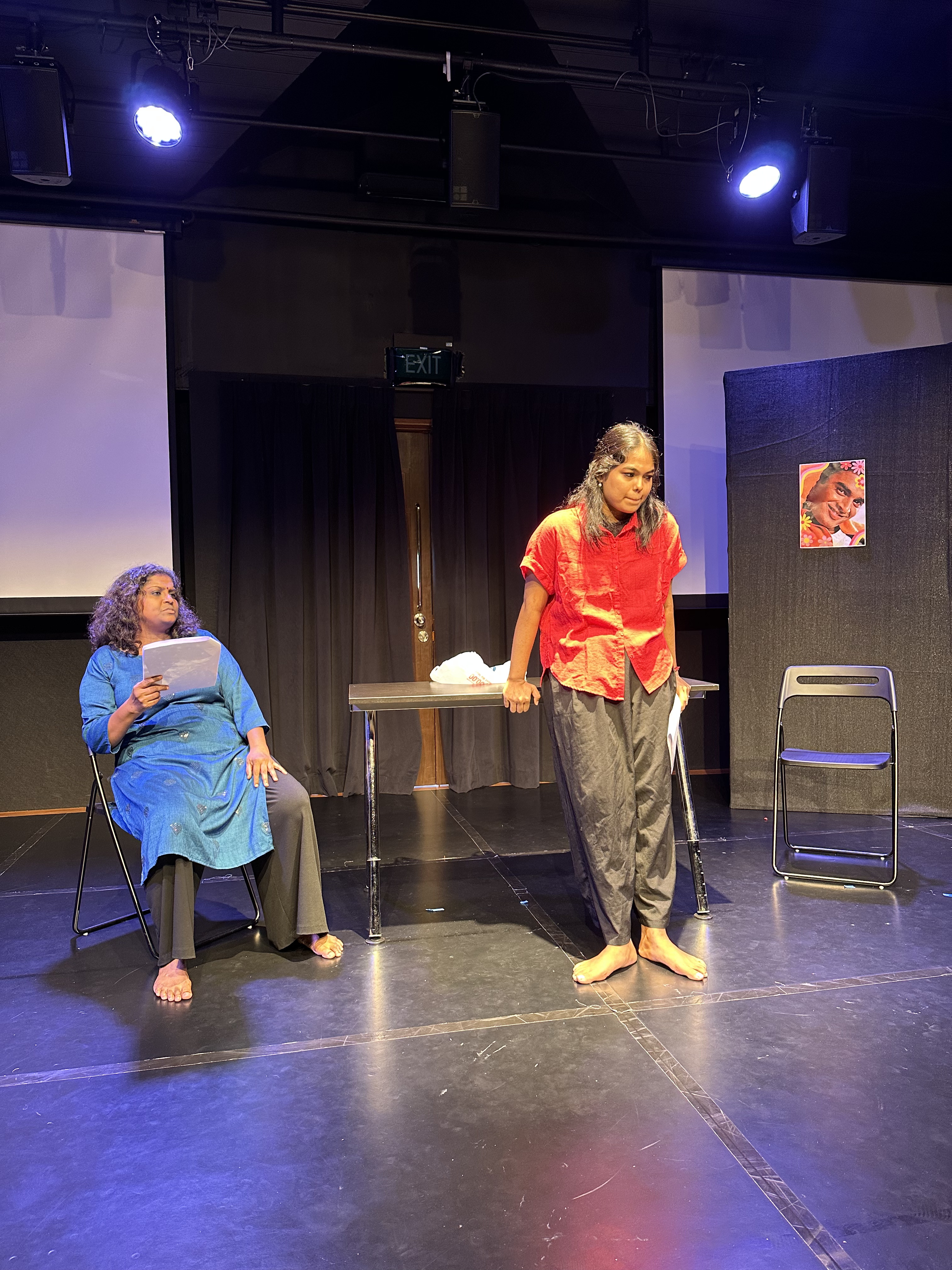 Two actresses on stage, one seated with a script in hand, and the other standing, hands on a table behind her.