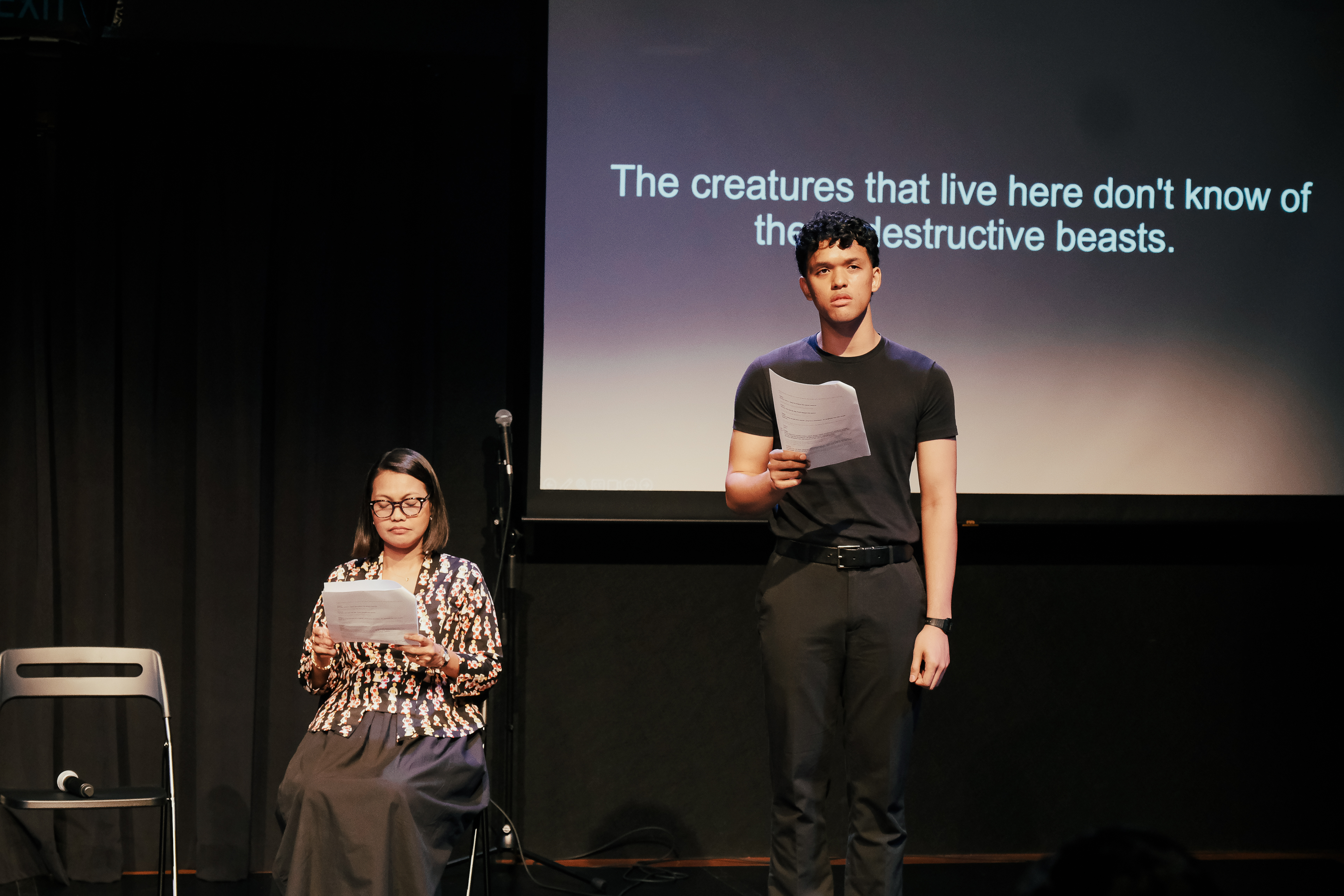 A photograph of actors Irsyad Dawood, standing, and Siti Hajar Abdul Gani, seated, reading from scripts with a projection of English surtitles behind them.