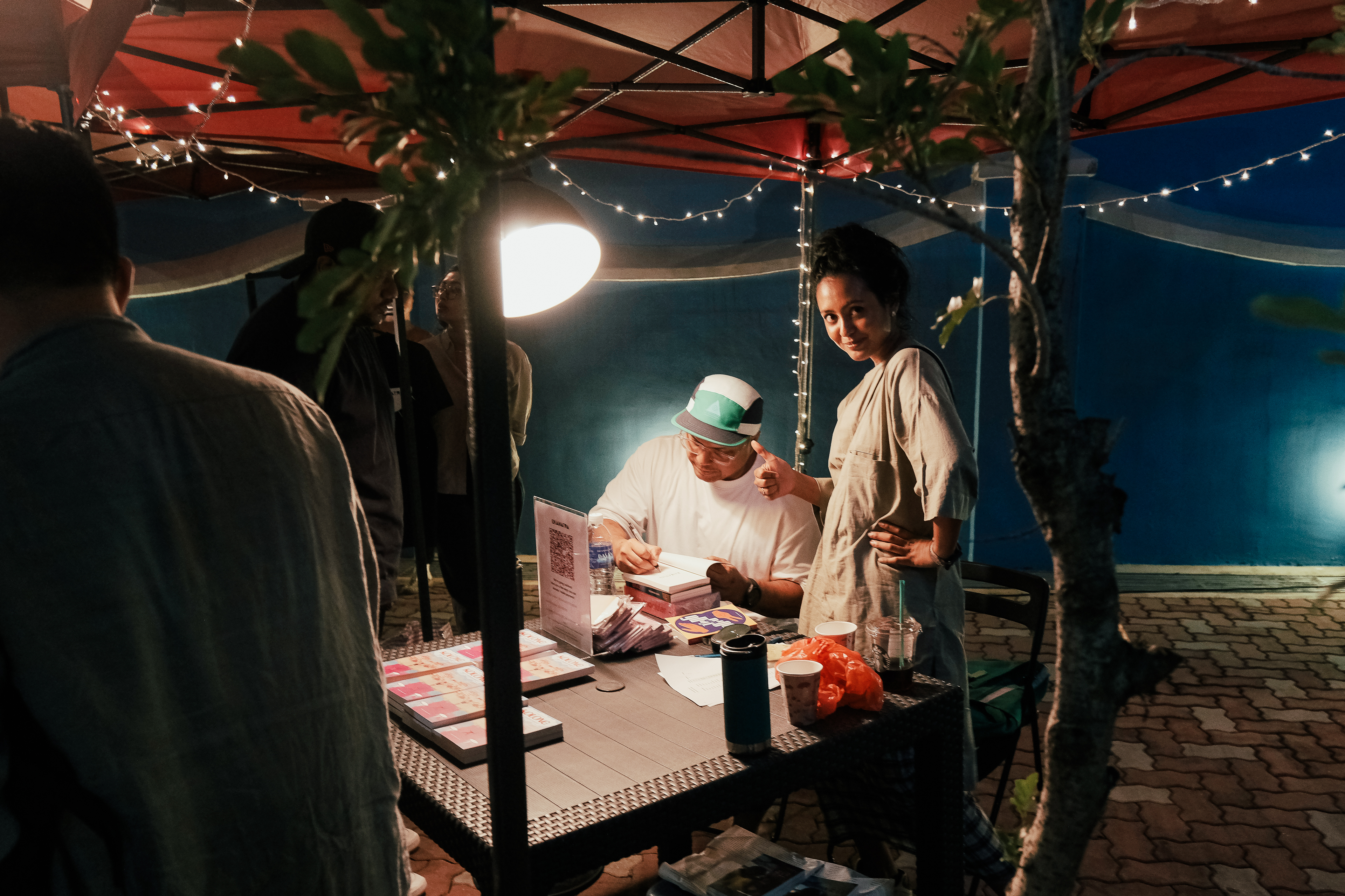 A photograph of Big signing his book, Kolong, seated at a table under a red gazebo.