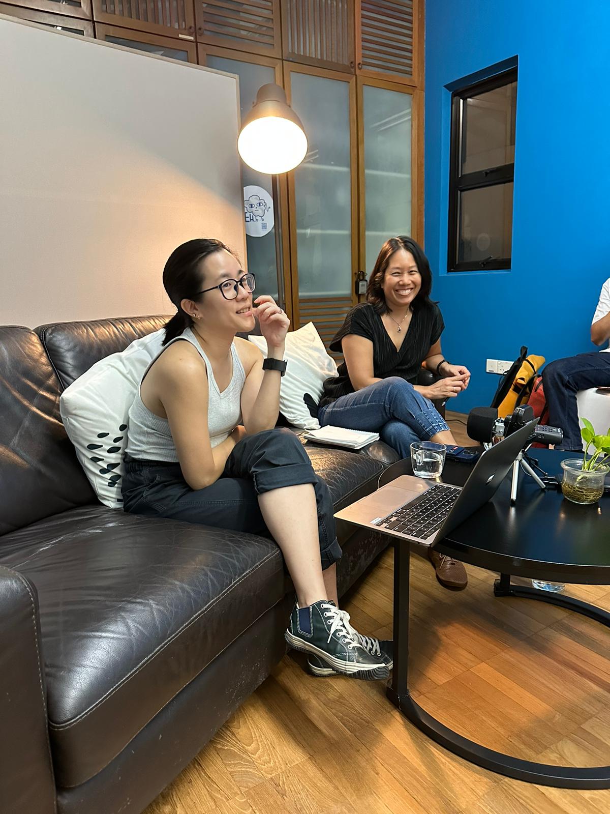 A photograph of Michelle Tan and Juliet Chia seated on a black couch with a laptop and a lounge table in front of them.