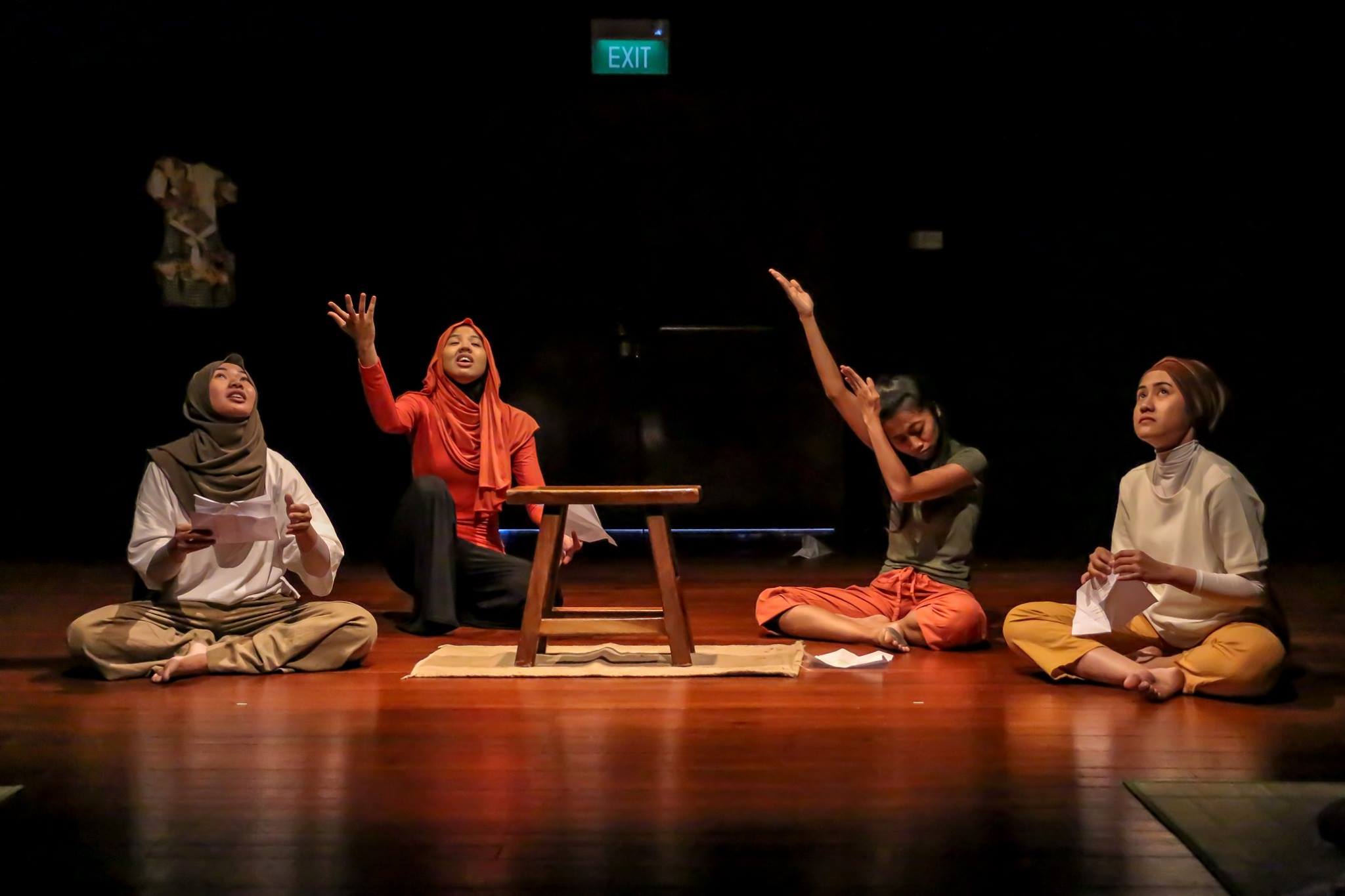 Four female-presenting persons seated on a wooden floor, gesturing and looking upwards.