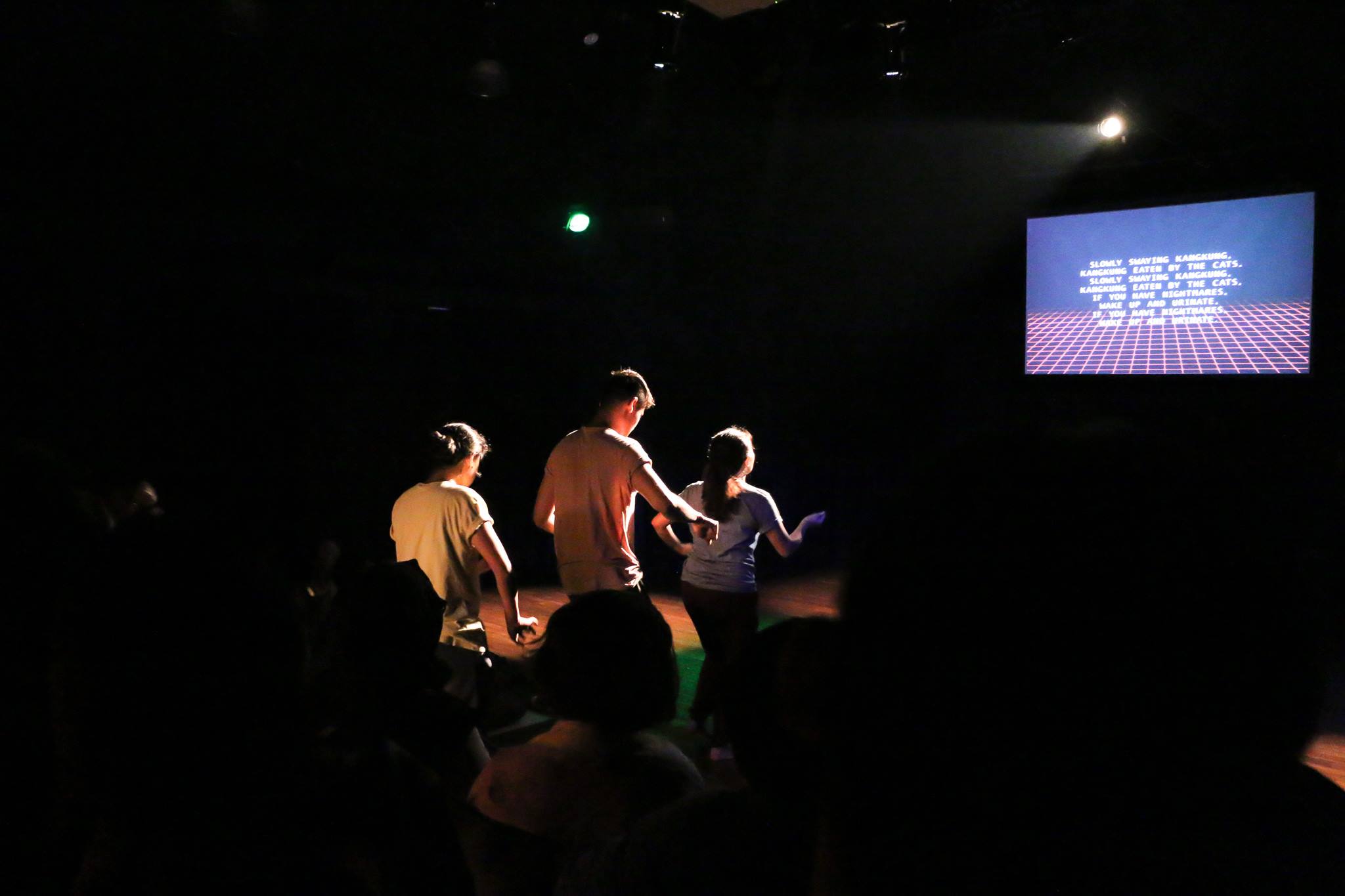 Three persons dancing while facing a projector screen displaying lyrics, karaoke-style.