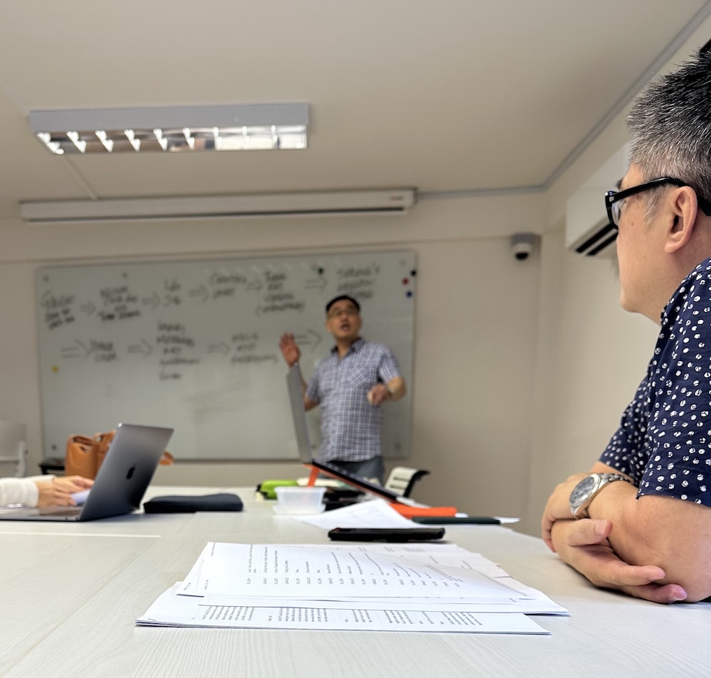 A male-presenting person presenting against a whiteboard, with another person watching him from a distance.