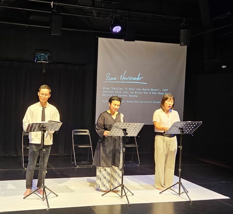 Three actors standing in a line on a white sheet, reading from music stands.