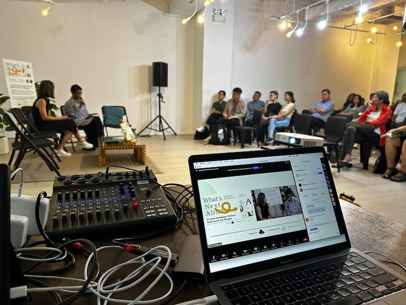 A laptop presenting on Zoom. In the background are two speakers speaking to an audience.