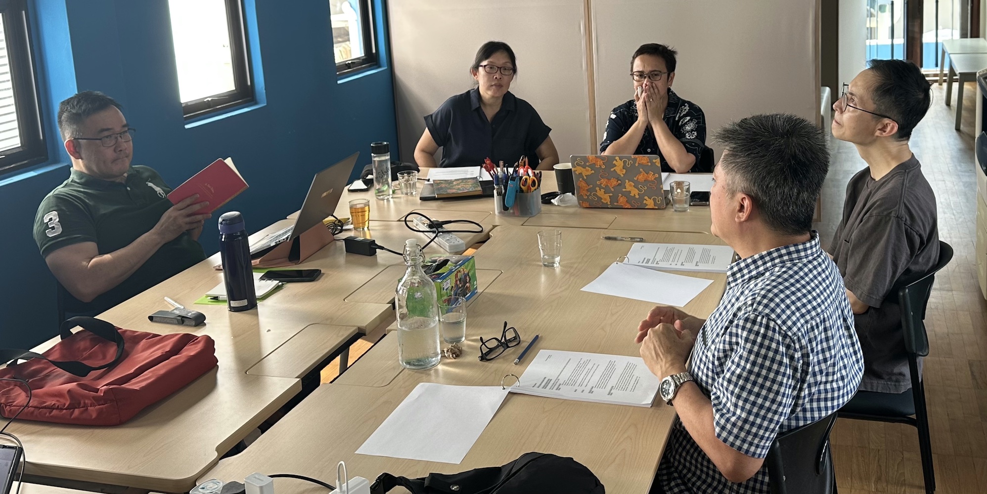 Five persons seated around some tables, reading from scripts.