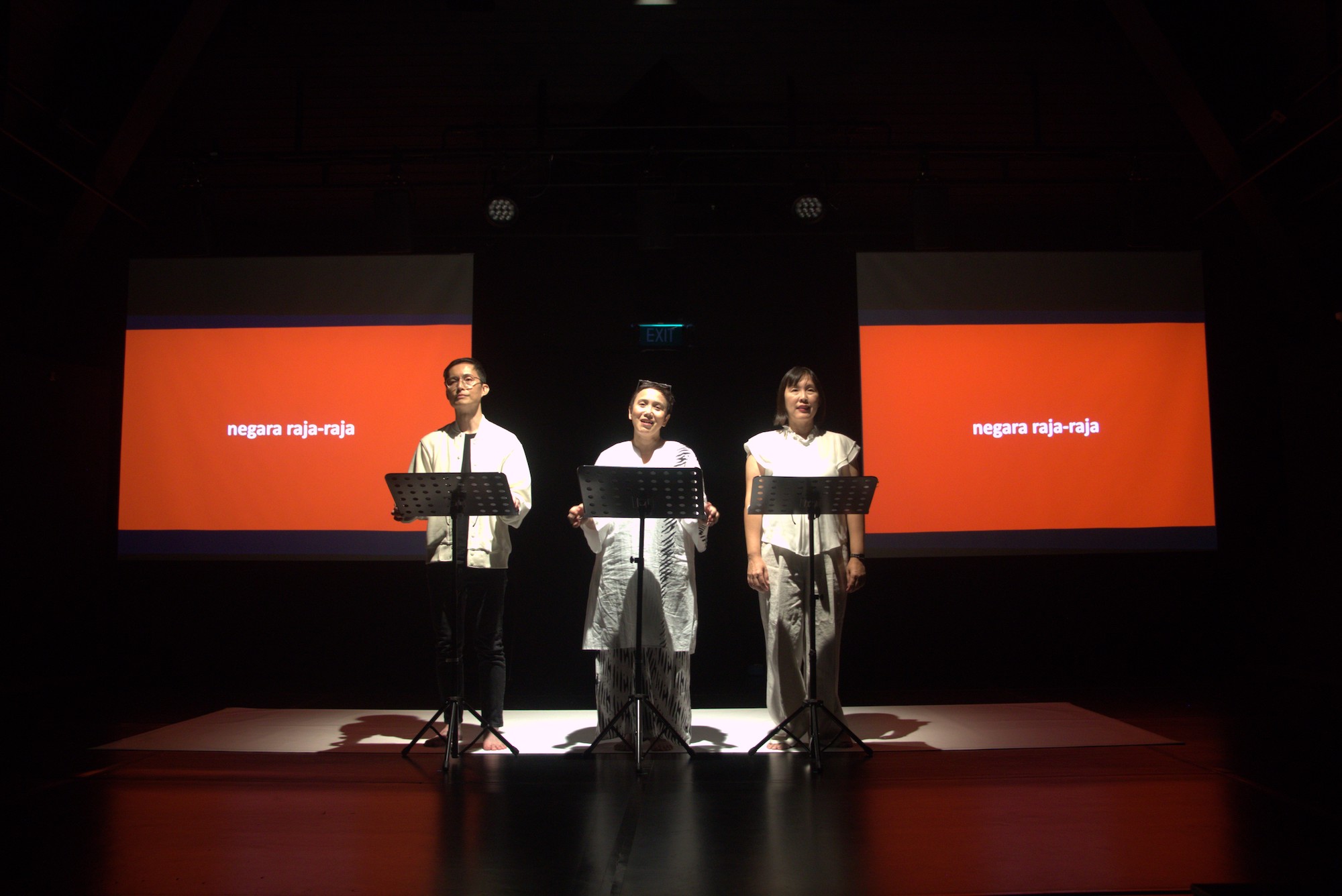 Three performers in white clothing standing behind three music stands. Behind them are two projector screens with red backgrounds.