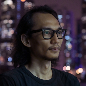 Headshot of Ridhwan Saidi, wearing black glasses, framed by a blurry cityscape background.