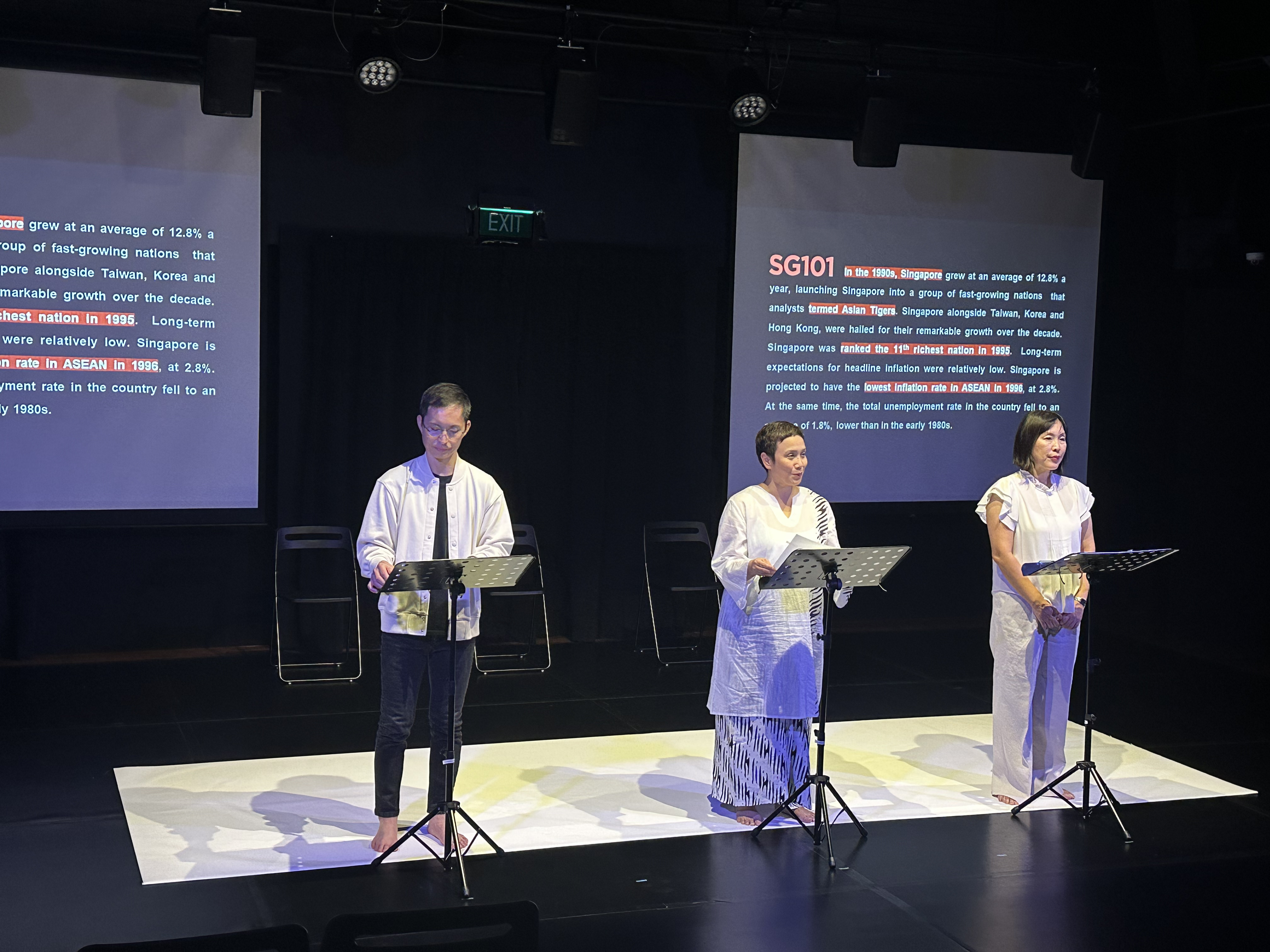 Three actors standing in front of music stands against the backdrop of two screens.