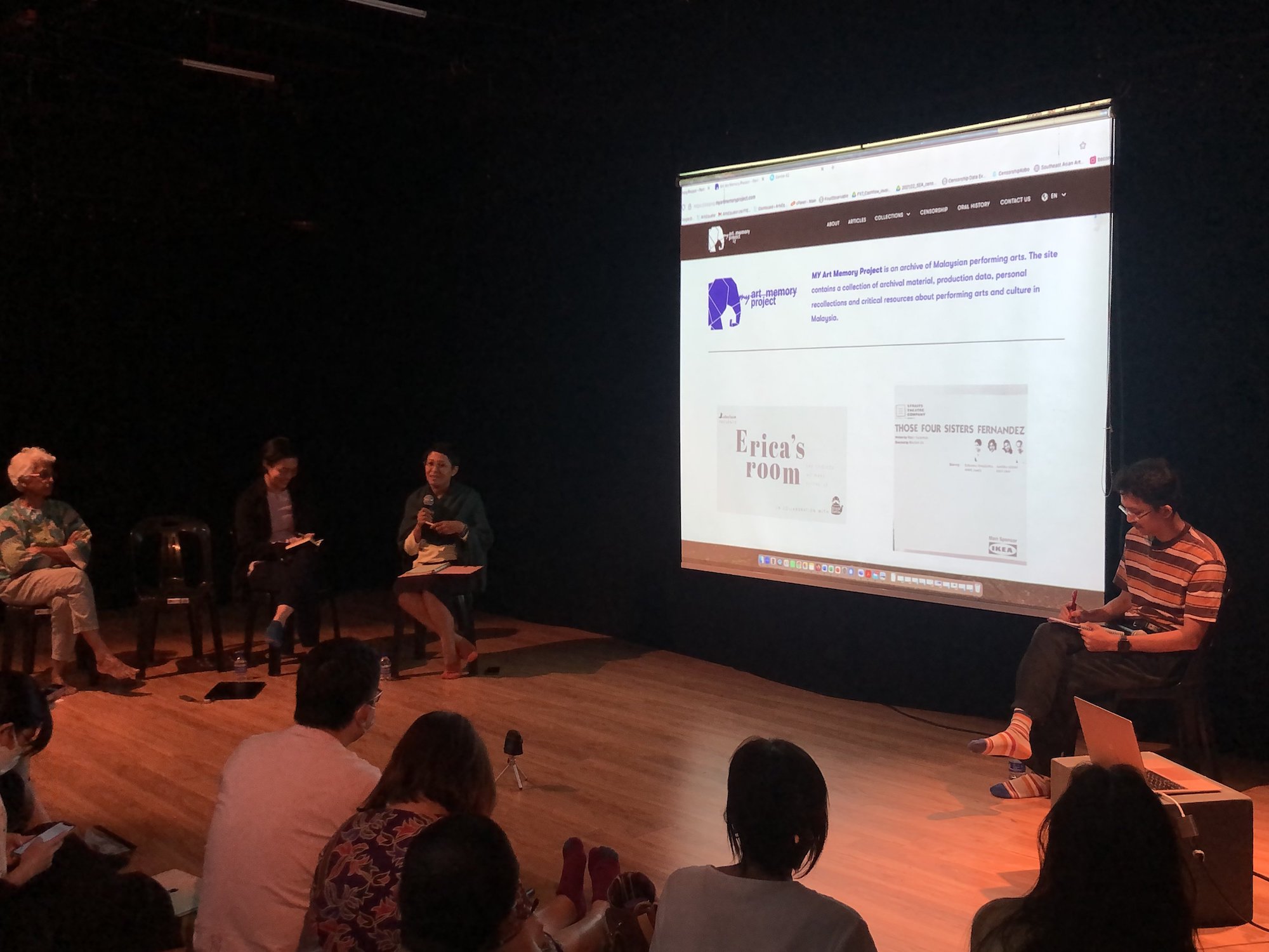 Four persons seated around a projector screen displaying a webpage. One is speaking into a microphone.