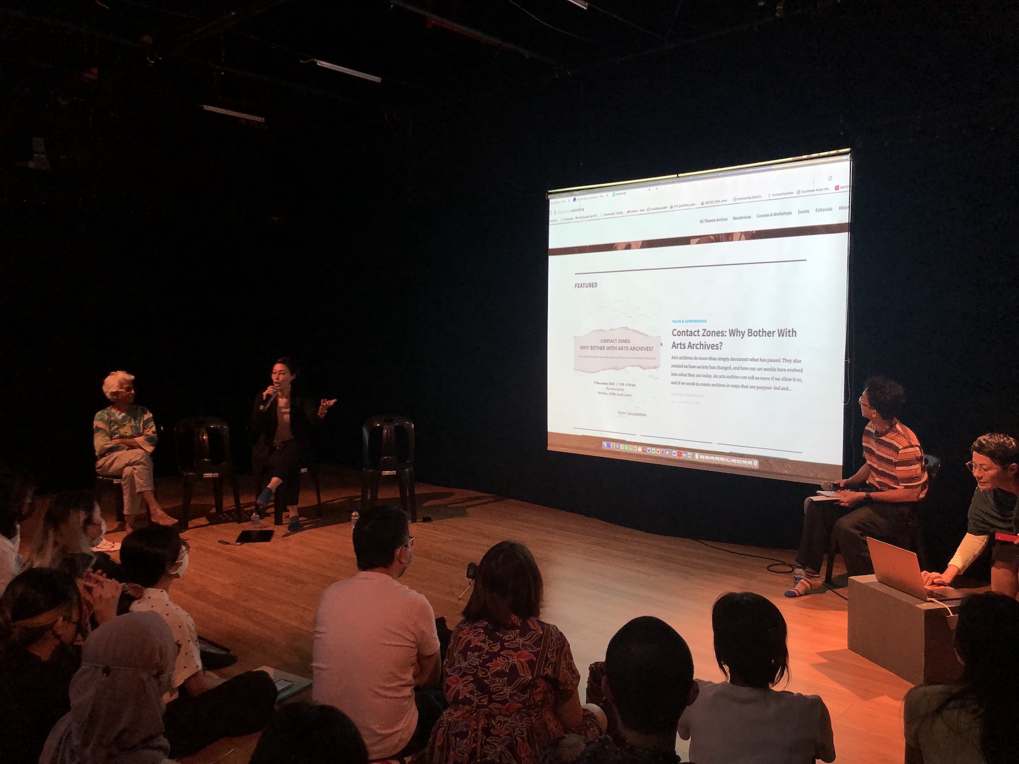 Four persons sat around a projector screen, one of them speaking into a microphone and gesturing to the screen.