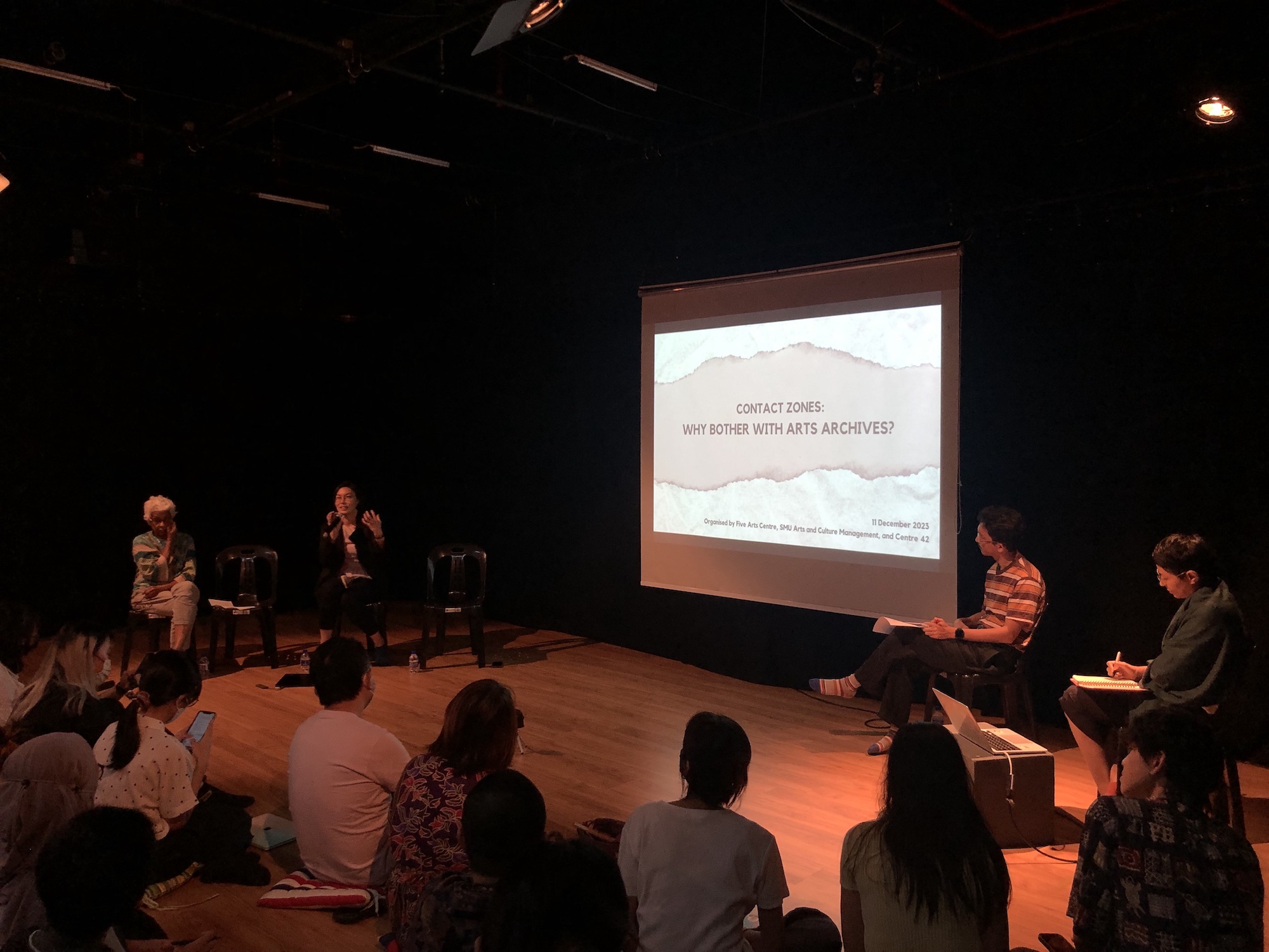 Four persons seated around a projector screen. On the projector is a slide that says 'Contact Zones: Why Bother With Arts Archives?'