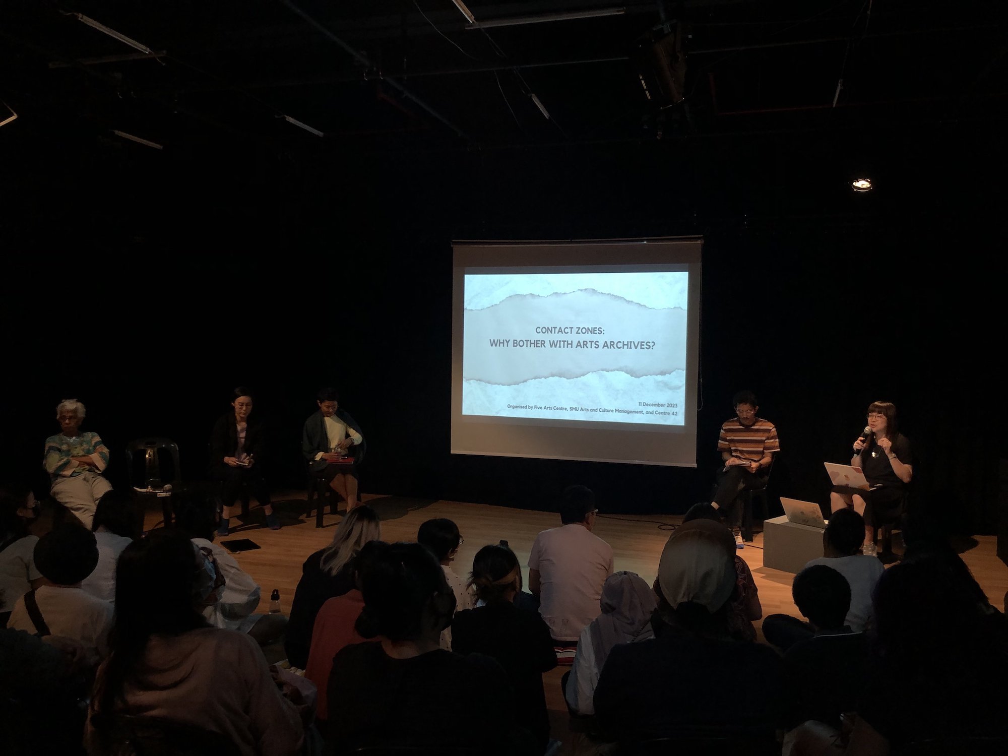 Four persons seated around a projector screen. The rightmost person is holding a microphone and speaking into it.