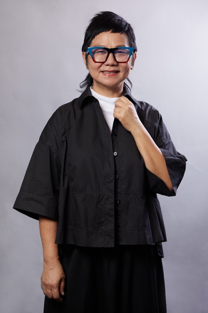 A half-body shot of Jalyn Han, wearing a black jacket and white top and glasses, smiling, holding on to her collar.